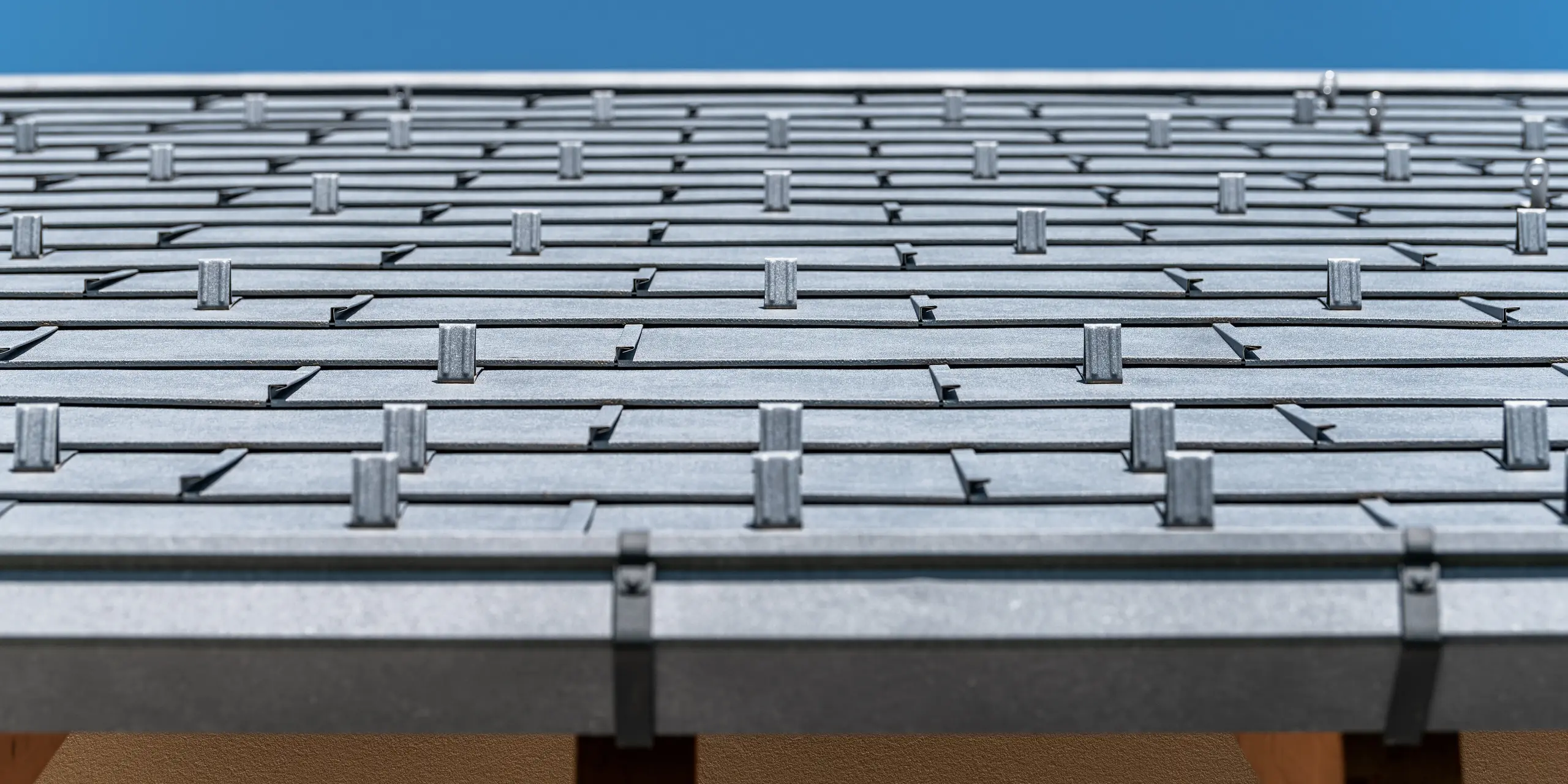 Detailný pohľad na strechu PREFA pokrytú strešným panelom R.16 vo farbe P.10 antracit. Na obrázku sú precízne spracované hliníkové strešné panely. Záber vyzdvihuje nápadnú štruktúru a moderný dizajn strechy, ktorý kontrastuje s jasne modrou oblohou a podčiarkuje inovatívne a odolné riešenie zastrešenia PREFA.