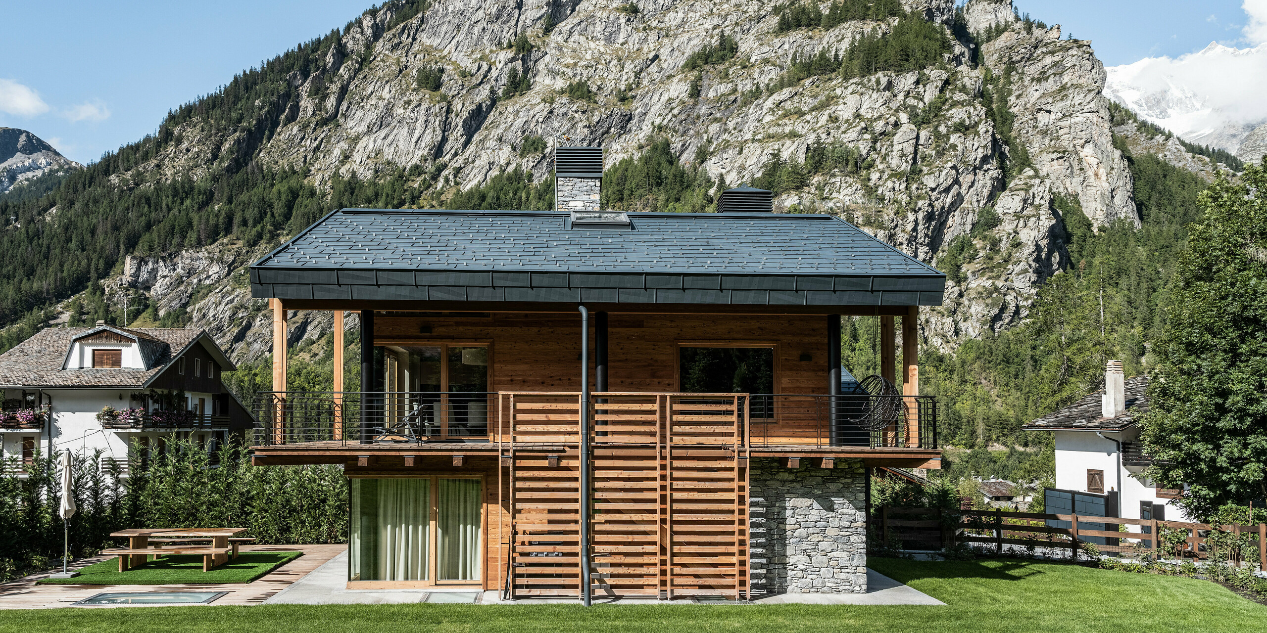 Idylická vila v Courmayeur s harmonickou kombináciou dreva, kameňa a hliníka na fasáde. Strešné a fasádne panely PREFA FX.12 v P.10 kamenná šedá sú prezentované na pôsobivom horskom pozadí, ktoré zdôrazňuje alpskú atmosféru. Výrazné detaily, ako je priestranné zábradlie balkóna a veľké okná, poskytujú otvorenú a prívetivú atmosféru, zatiaľ čo udržiavaná záhrada pozýva na relaxáciu vonku.