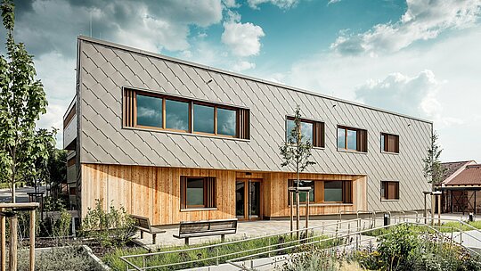 Čelný pohľad na detské zariadenie v Alzenau zobrazuje pozývajúci hlavný vstup s harmonickou kombináciou bronzovo sfarbených PREFA fasádnych šablón 44 × 44 a vertikálnej drevenej fasády na prízemí. Jasne štruktúrovaný hliníkový obklad vytvára elegantný šupinový vzor, ktorý je rytmicky prerušovaný veľkorysými oknami s drevenými rámami a vertikálnymi lamelami. V popredí bezbariérové komunikačné trasy, kvetinové záhony a lavičky podčiarkujú vysokú kvalitu pobytového priestoru. Súhra trvácneho hliníka, prírodného dreva a premyslene riešených exteriérových plôch robí z budovy architektonický vzorový projekt udržateľných, funkčných a esteticky hodnotných vzdelávacích stavieb s fasádnymi systémami PREFA.