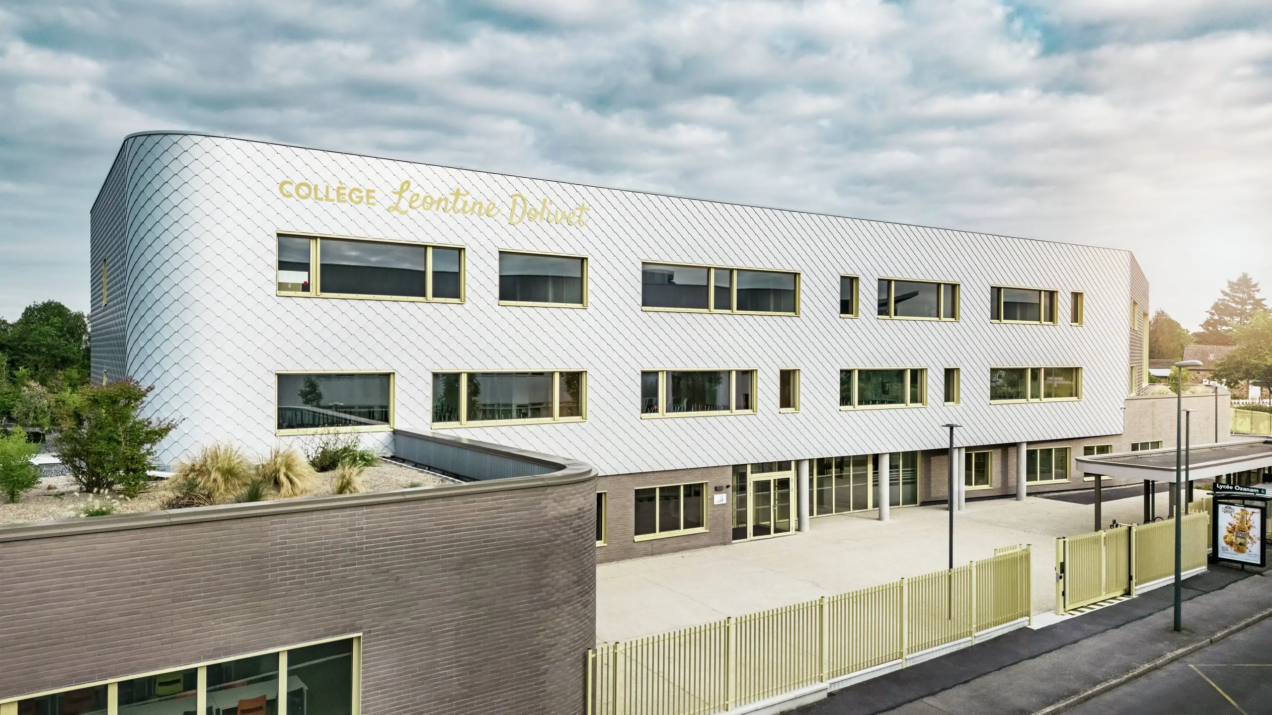 Pohľad na fasádu školy Collège Léontine Dolivet v Cesson-Sévigné s modernou hliníkovou fasádou z PREFA fasádnych šablón v striebornom metalickom vyhotovení. Meniaci sa povrch dodáva budove ľahkosť, zatiaľ čo čisté línie a veľké okná zaisťujú transparentnosť. Žlté okenné rámy dodávajú budove živý akcent. V popredí je vidieť svetlé predsieň, plot a susednú ulicu. Trvanlivé fasádne riešenie PREFA kombinuje dizajn, odolnosť voči poveternostným vplyvom a udržateľnosť – architektonický pohľad upútavajúci elegantným dizajnom.
