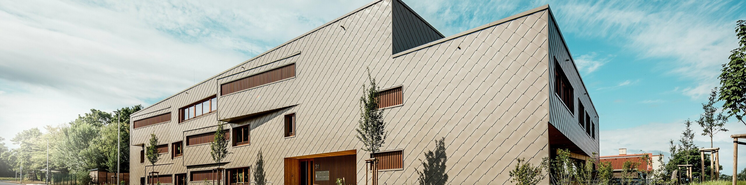 Z výraznej perspektívy zospodu sa detské jasle v Alzenau prezentujú svojimi výraznými, dynamickými líniami a vysokokvalitnou hliníkovou fasádou PREFA v bronzovej farbe. Diagonálne usporiadanie šablón 44 × 44 dodáva budove jasnú štruktúru a elegantný, homogénny povrch. Posunuté okenné otvory a vertikálne drevené lamely dodávajú rytmické akcenty, zatiaľ čo minimalistický dizajn signalizuje modernú zdržanlivosť. Kombinácia odolného hliníka, prírodného dreva a premysleného konceptu zelene pozdĺž ulice podčiarkuje vysoké nároky na udržateľnosť a kvalitu stavby. Tento obrázok pôsobivo ukazuje, ako môže funkčná architektúra s produktmi PREFA presvedčiť svojím dizajnom a stanoviť nové štandardy v urbanistike.