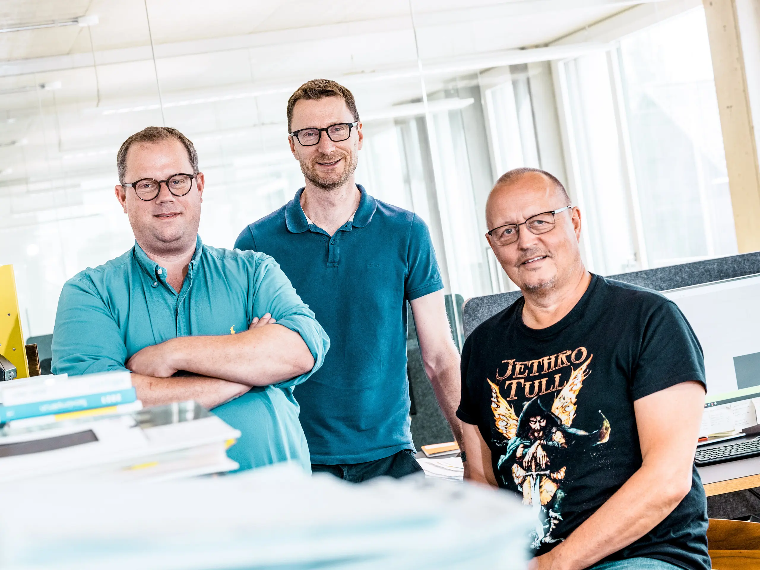  Tímová fotografia architektov Kaundbe v ich modernej kancelárii: Traja architekti stoja sebavedome pred svojimi pracoviskami a technickými dokumentmi. V popredí ležia stohy plánov a projektové zložky, v pozadí sú vidieť počítačové monitory s konštrukčnými výkresmi. Scéna vyžaruje kompetentnosť, spoluprácu a technickú precíznosť – kľúčové hodnoty pri plánovaní a realizácii fasádnych riešení s hliníkovými produktmi PREFA.