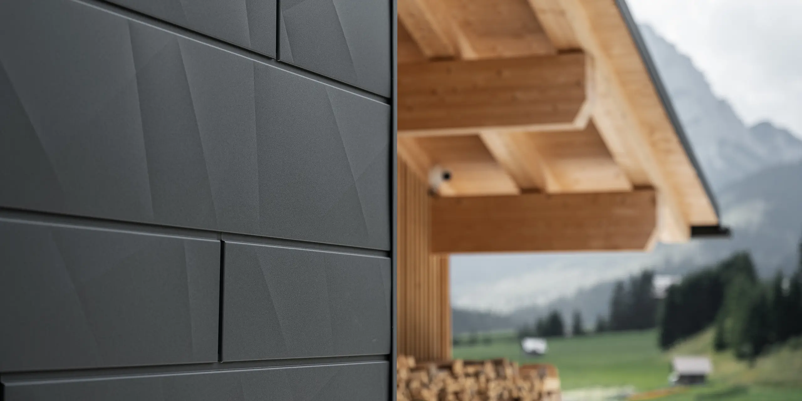 Detailný záber na fasádne panely Siding.X v čiernej farbe P.10 od spoločnosti PREFA na budove Casa Hoffe v Sappade v Taliansku. Hliníkový fasádny systém je šikovne zakomponovaný do moderného, abstraktného dizajnu, ktorý vytvára príjemný kontrast s viditeľnými drevenými prvkami krovu. Toto inovatívne riešenie fasády sa dokonale začleňuje do alpského prostredia a predstavuje spojenie funkčnosti, trvácnosti a estetického stavebného štýlu.