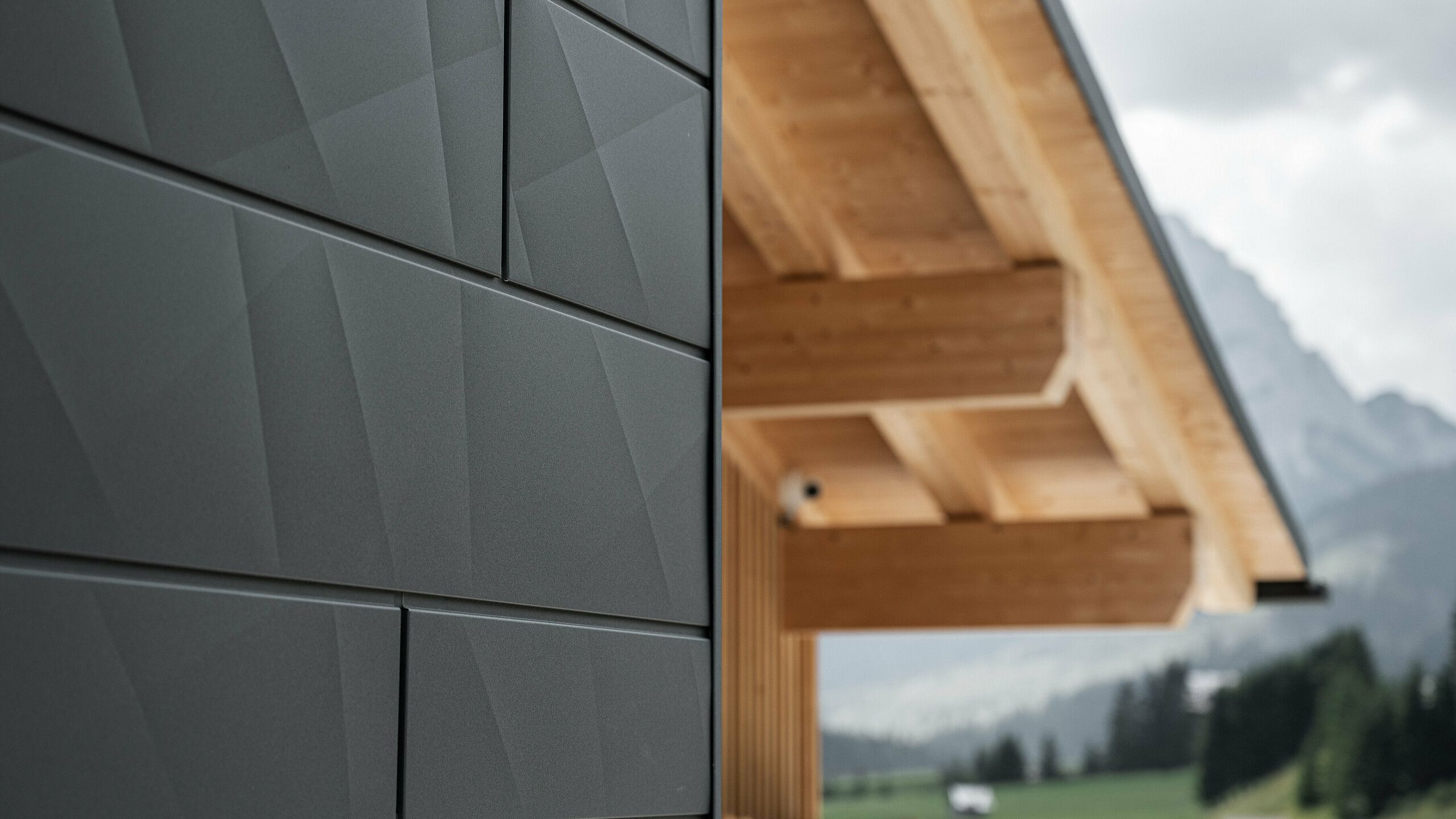Detailný záber na fasádne panely Siding.X vo farbe P.10 čierna v Casa Hoffe v Sappada, Taliansko. Hliníkový fasádny systém je šikovne inštalovaný v modernom, abstraktnom dizajne, ktorý vytvára príťažlivý kontrast k viditeľným dreveným prvkom strešnej konštrukcie. Inovatívne riešenie fasády bez problémov zapadá do alpského prostredia a predstavuje kombináciu funkčnosti, odolnosti a estetickej stavebnej kultúry.