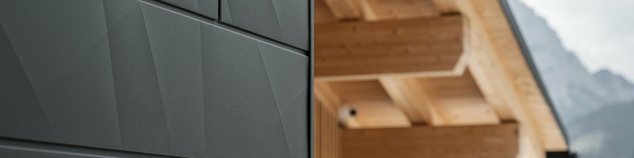Detailný záber na fasádne panely Siding.X vo farbe P.10 čierna v Casa Hoffe v Sappada, Taliansko. Hliníkový fasádny systém je šikovne inštalovaný v modernom, abstraktnom dizajne, ktorý vytvára príťažlivý kontrast k viditeľným dreveným prvkom strešnej konštrukcie. Inovatívne riešenie fasády bez problémov zapadá do alpského prostredia a predstavuje kombináciu funkčnosti, odolnosti a estetickej stavebnej kultúry.