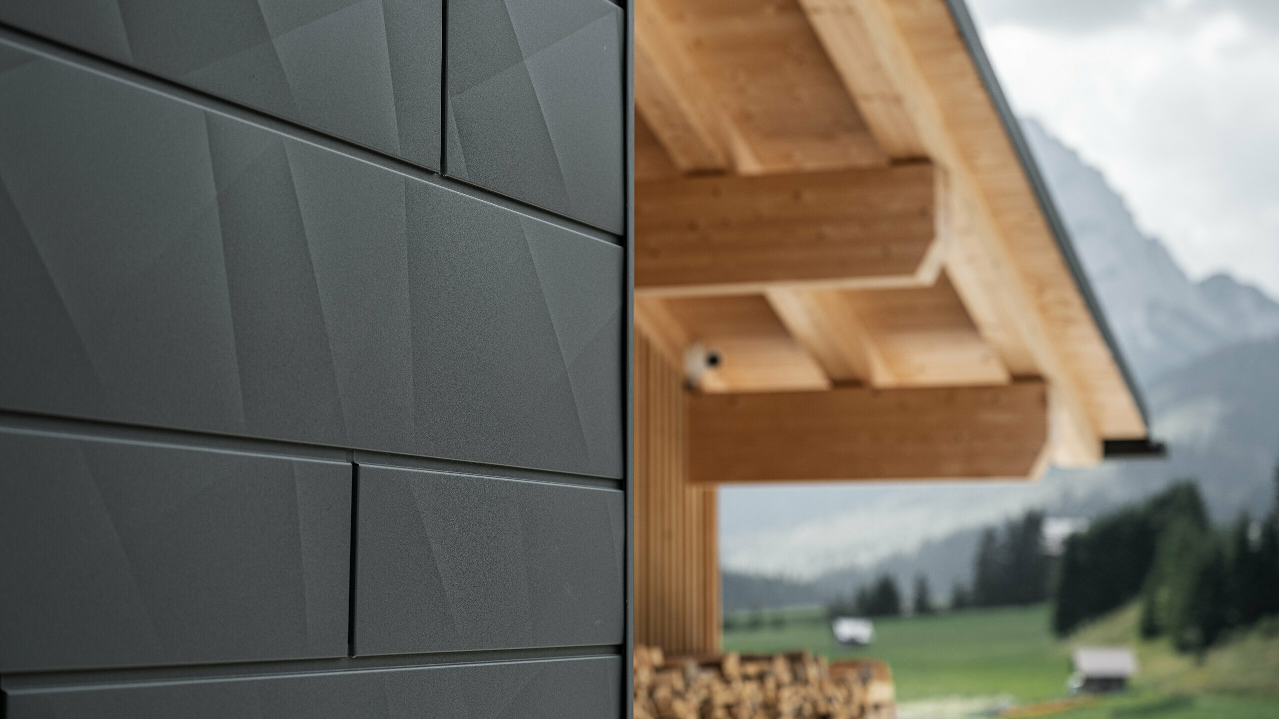 Detailný záber na fasádne panely Siding.X vo farbe P.10 čierna v Casa Hoffe v Sappada, Taliansko. Hliníkový fasádny systém je šikovne inštalovaný v modernom, abstraktnom dizajne, ktorý vytvára príťažlivý kontrast k viditeľným dreveným prvkom strešnej konštrukcie. Inovatívne riešenie fasády bez problémov zapadá do alpského prostredia a predstavuje kombináciu funkčnosti, odolnosti a estetickej stavebnej kultúry.