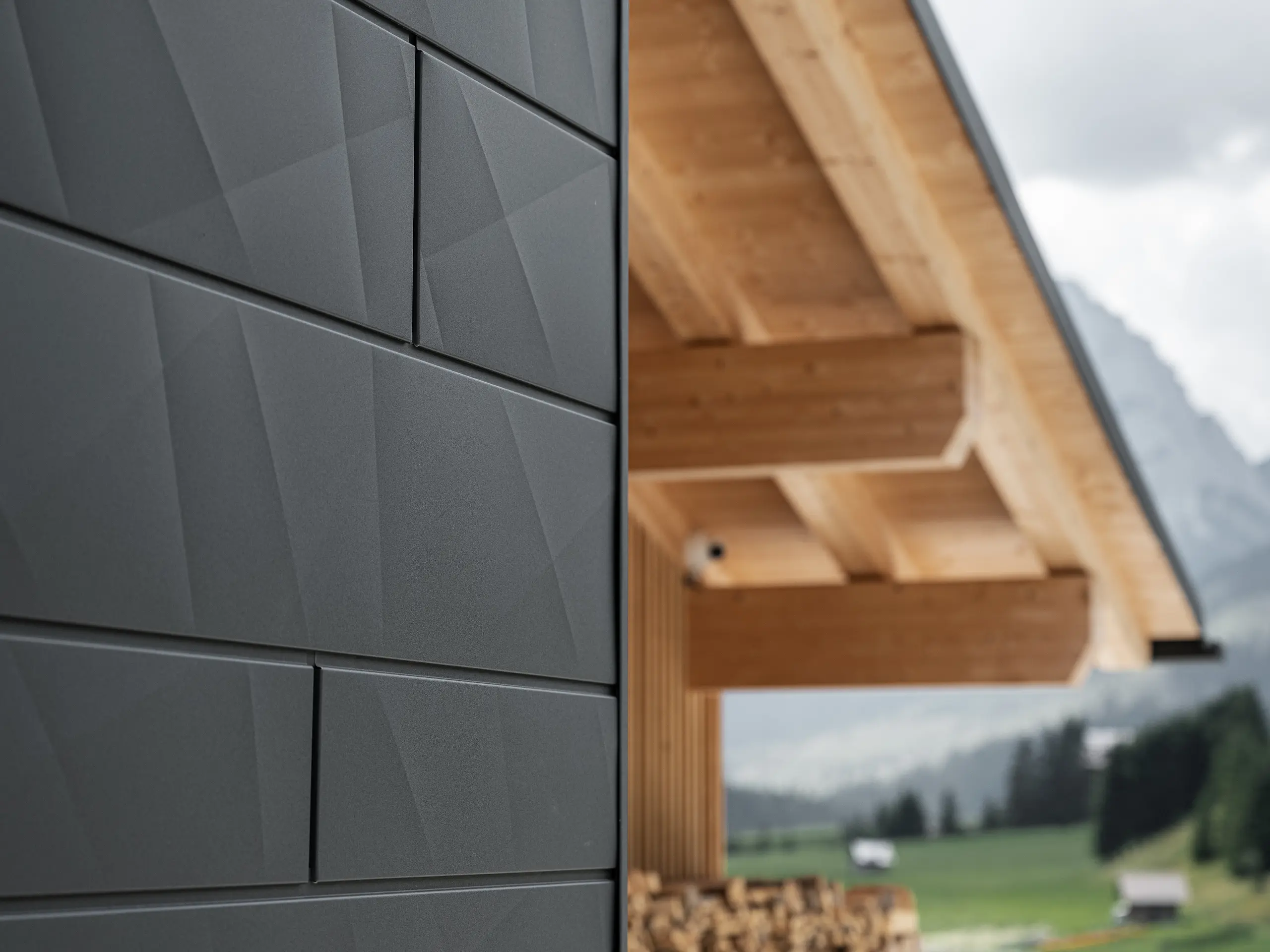 Detailný záber na fasádne panely Siding.X v čiernej farbe P.10 od spoločnosti PREFA na budove Casa Hoffe v Sappade v Taliansku. Hliníkový fasádny systém je šikovne zakomponovaný do moderného, abstraktného dizajnu, ktorý vytvára príjemný kontrast s viditeľnými drevenými prvkami krovu. Toto inovatívne riešenie fasády sa dokonale začleňuje do alpského prostredia a predstavuje spojenie funkčnosti, trvácnosti a estetického stavebného štýlu.