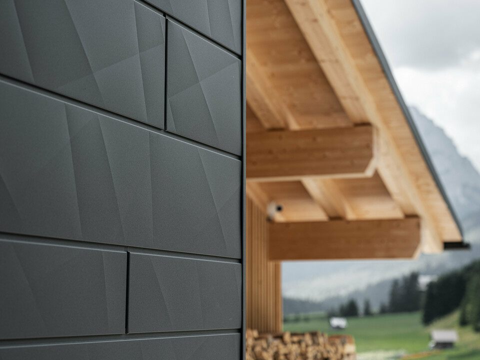 Detailný záber na fasádne panely Siding.X vo farbe P.10 čierna v Casa Hoffe v Sappada, Taliansko. Hliníkový fasádny systém je šikovne inštalovaný v modernom, abstraktnom dizajne, ktorý vytvára príťažlivý kontrast k viditeľným dreveným prvkom strešnej konštrukcie. Inovatívne riešenie fasády bez problémov zapadá do alpského prostredia a predstavuje kombináciu funkčnosti, odolnosti a estetickej stavebnej kultúry.