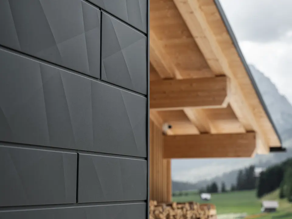 Detailný záber na fasádne panely Siding.X v čiernej farbe P.10 od spoločnosti PREFA na budove Casa Hoffe v Sappade v Taliansku. Hliníkový fasádny systém je šikovne zakomponovaný do moderného, abstraktného dizajnu, ktorý vytvára príjemný kontrast s viditeľnými drevenými prvkami krovu. Toto inovatívne riešenie fasády sa dokonale začleňuje do alpského prostredia a predstavuje spojenie funkčnosti, trvácnosti a estetického stavebného štýlu.