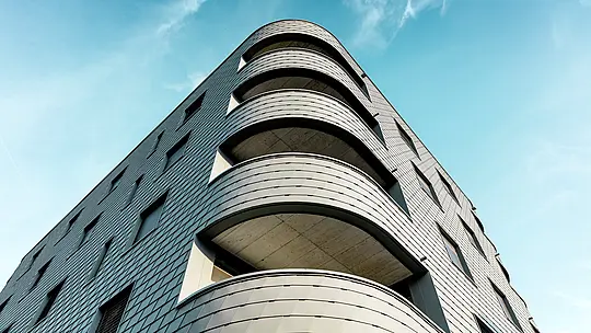 Dynamický pohľad na spodnú časť modernej obytnej a obchodnej budovy s výrazne zaobleným rohom a plynulými balkónovými pásmi. Šupinová fasáda z PREFA fasádnych šindľov v odtieni P.10 svetlošedá pokrýva budovu ako precízne spracovaná hliníková koža a zdôrazňuje jej jemné polomery. Homogénny povrch podčiarkuje čisté línie, zatiaľ čo ľahký, odolný voči poveternostným vplyvom hliník PREFA zaručuje aj pri silných zaobleniach najvyššiu tvarovú stabilitu, dlhú životnosť a nízke nároky na údržbu – architektonický pohľad upútavajúci pred žiarivo modrou oblohou.