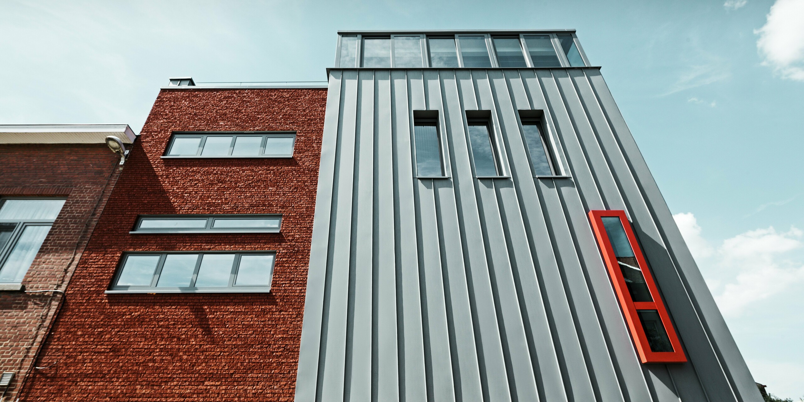 Štýlová kombinácia tradičnej tehlovej fasády a inovatívnej hliníkovej fasády PREFALZ v patinovo šedej farbe. Výrazná vertikálna štruktúra stojatých falcov sa snúbi s výraznými červenými oknami a čistými líniami okien. Odolný hliník trvalo chráni pred vplyvmi počasia a dodáva budove nadčasový moderný charakter.