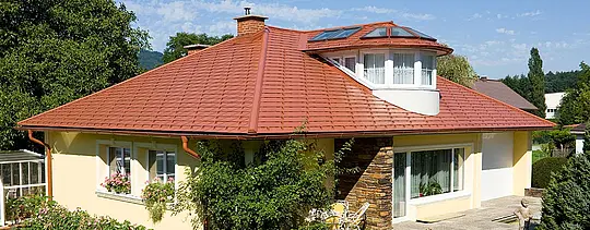 Rodinný dom s valbovou strechou a vikierom zastrešený hliníkovým šindľom PREFA vo vzhľade škridly v tehlovočervenej farbe.