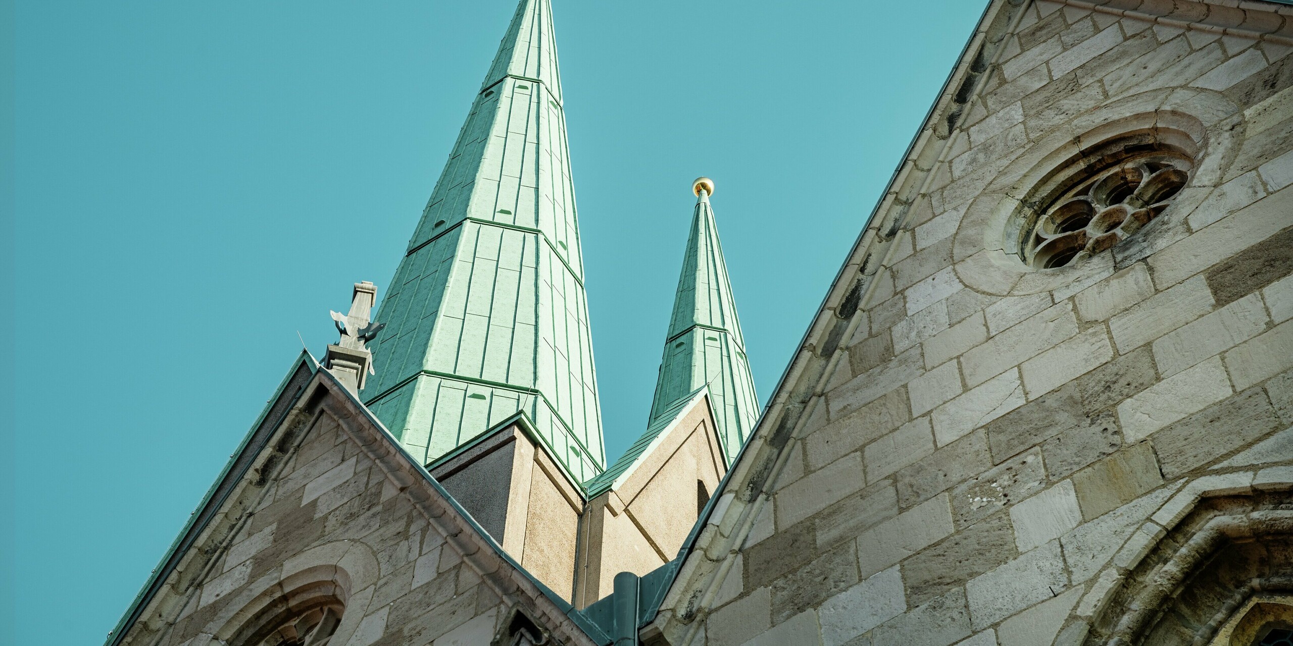 Stimmungsvolle Perspektive auf die eindrucksvoll sanierten Turmspitzen der Stadtkirche St. Johannis in Ellrich. Die schlanken, mit PREFALZ in P.10 Patinagrün eingedeckten Dachflächen aus Aluminium ragen elegant in den Himmel und setzen starke architektonische Akzente. Das hochwertige Blechdach ist wartungsfrei, extrem langlebig und ideal für historische Bauwerke mit moderner Nutzung.