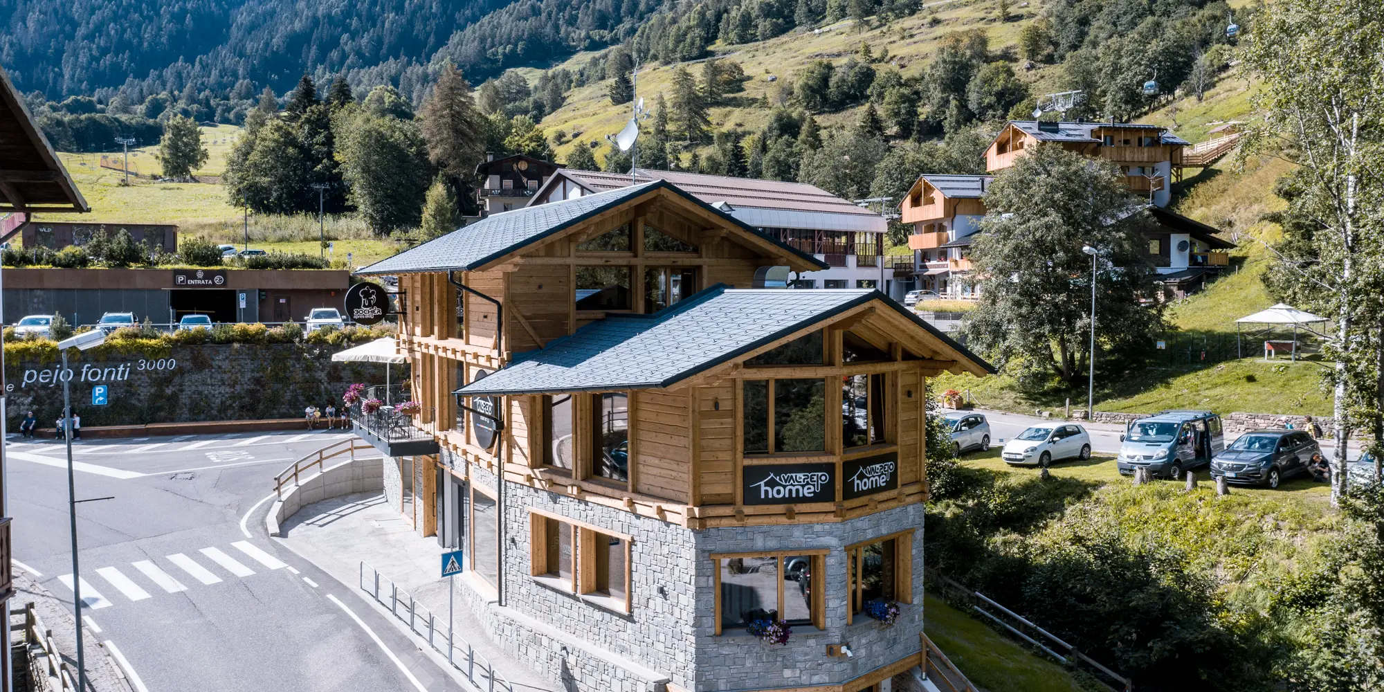 Pohľad na moderný après-ski bar v tradičnej drevenej architektúre s kamenno-sivými strešnými šindľami PREFA DS.19, vyrobenými z hliníka, začlenený do idylickej horskej krajiny so sviežou zeleňou a výhľadom na Alpy.