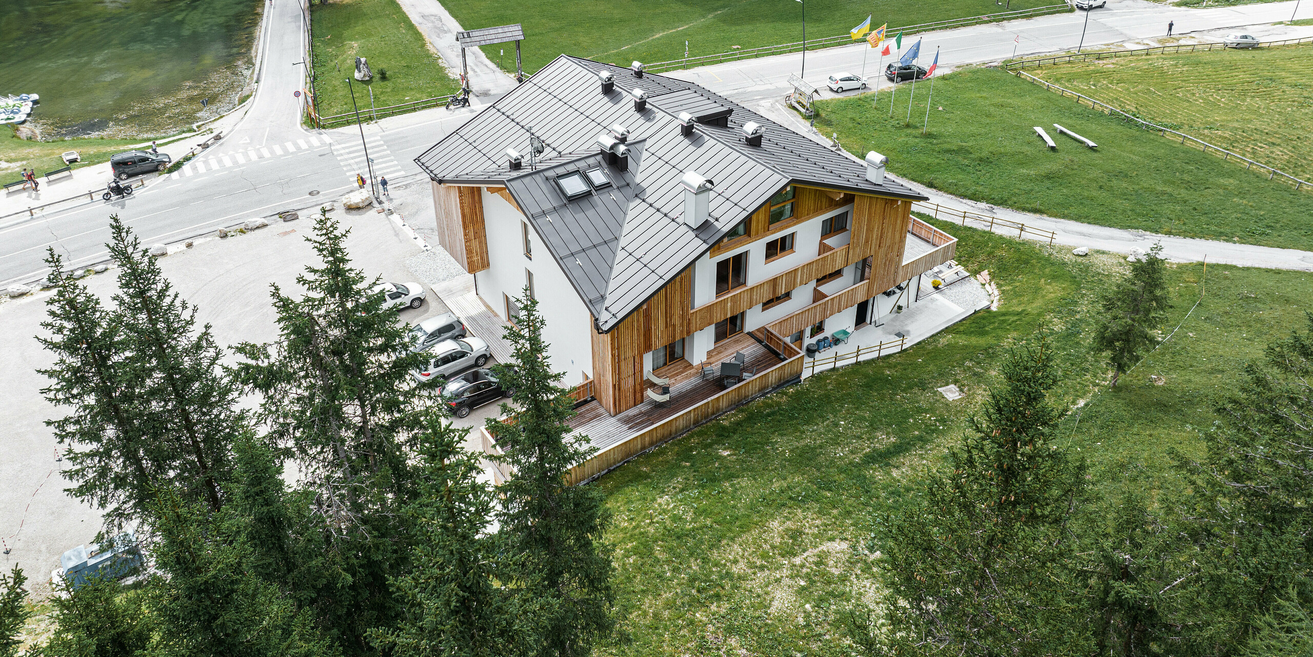 Letecký pohľad na chatu Alpenrose v Auronzo di Cadore, BL (Taliansko). Budova sa nachádza uprostred malebnej horskej krajiny, je vybavená strechou PREFALZ vo farbe P.10 orieškovohnedá. Moderná chata kombinuje tradičnú drevenú architektúru s robustným a odolným strešným systémom PREFA, ktorý sa ideálne hodí do prírodného prostredia a nachádza sa vedľa hotela a malého jazera.