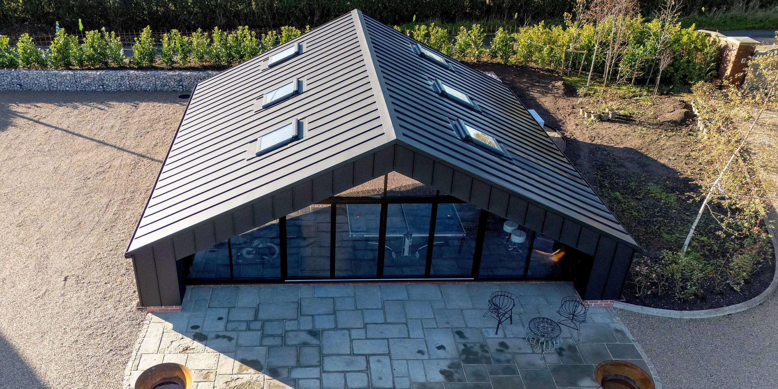 Vogelperspektive auf das moderne Freizeitgebäude mit vollständig verkleideter PREFA Aluminiumhülle und PREFALZ Dachfläche in P.10 Schwarz. Die klare, symmetrische Dachform, die großflächige Glasfront und der strukturierte Außenbereich erzeugen ein dynamisches, hochwertiges Erscheinungsbild. Die witterungsbeständige Aluminiumoberfläche garantiert langfristige Stabilität und architektonische Präzision.