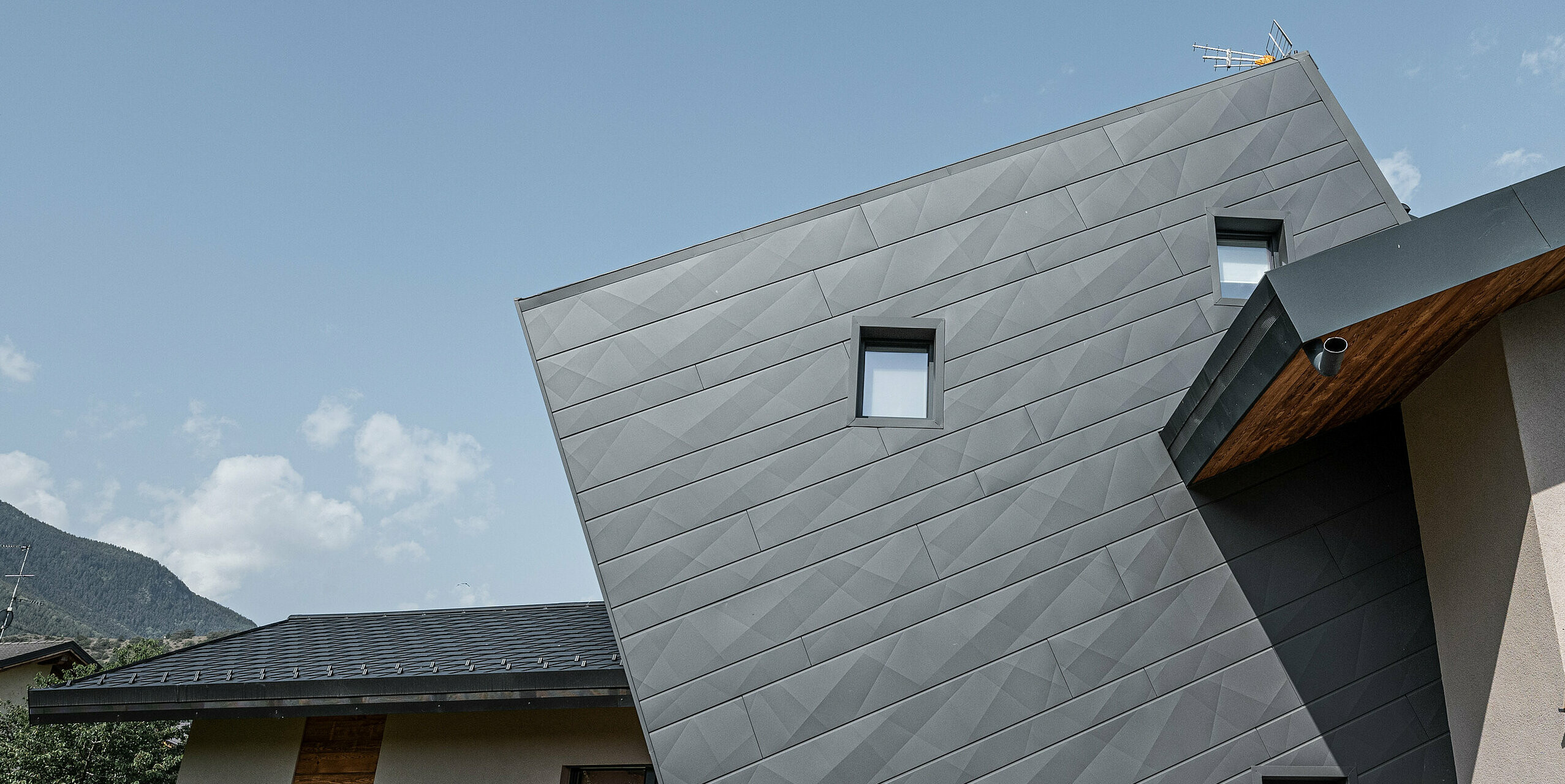 Vila Tia v talianskom Gressane má moderný dizajn fasády s obkladom PREFA Siding.X vo farbe P.10 tmavošedá. Dynamické usporiadanie panelov vytvára trojrozmerný vzhľad, ktorý je zdôraznený kontrastom s prírodnými drevenými prvkami a okolitou krajinou. Toto inovatívne špeciálne riešenie šikmých fasádnych panelov Siding.X je symbolom moderného a odolného dizajnu.