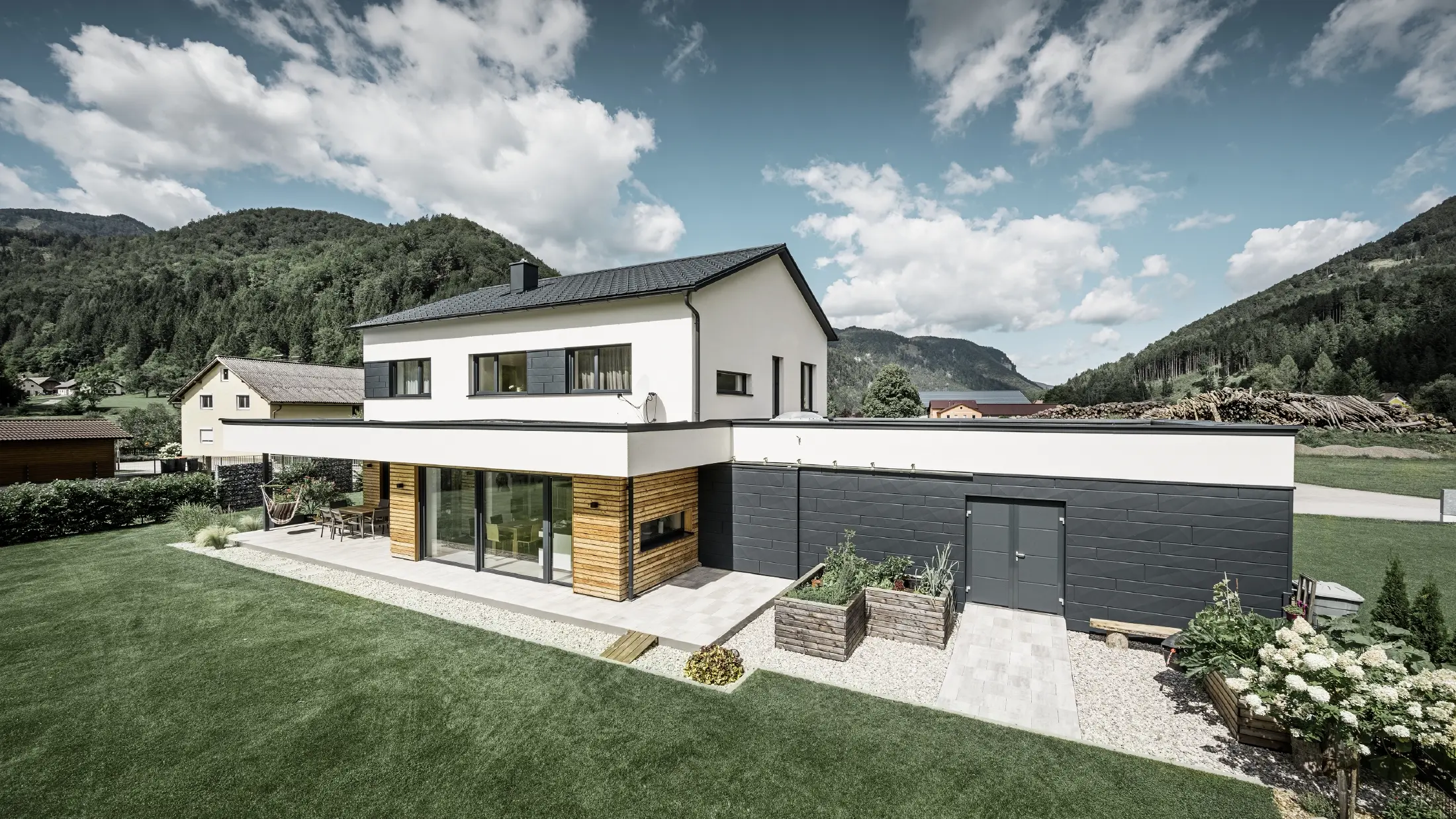 Moderný rodinný dom s bielou omietnutou fasádou a obkladom PREFA Siding.X v antracitovej farbe P.10, ktorý tvorí výrazný kontrast k prírodnému prostrediu. Krytá Falcovanou škridlou PREFA, odvodnenie strechy závesným žľabom PREFA a odtokovou rúrou.