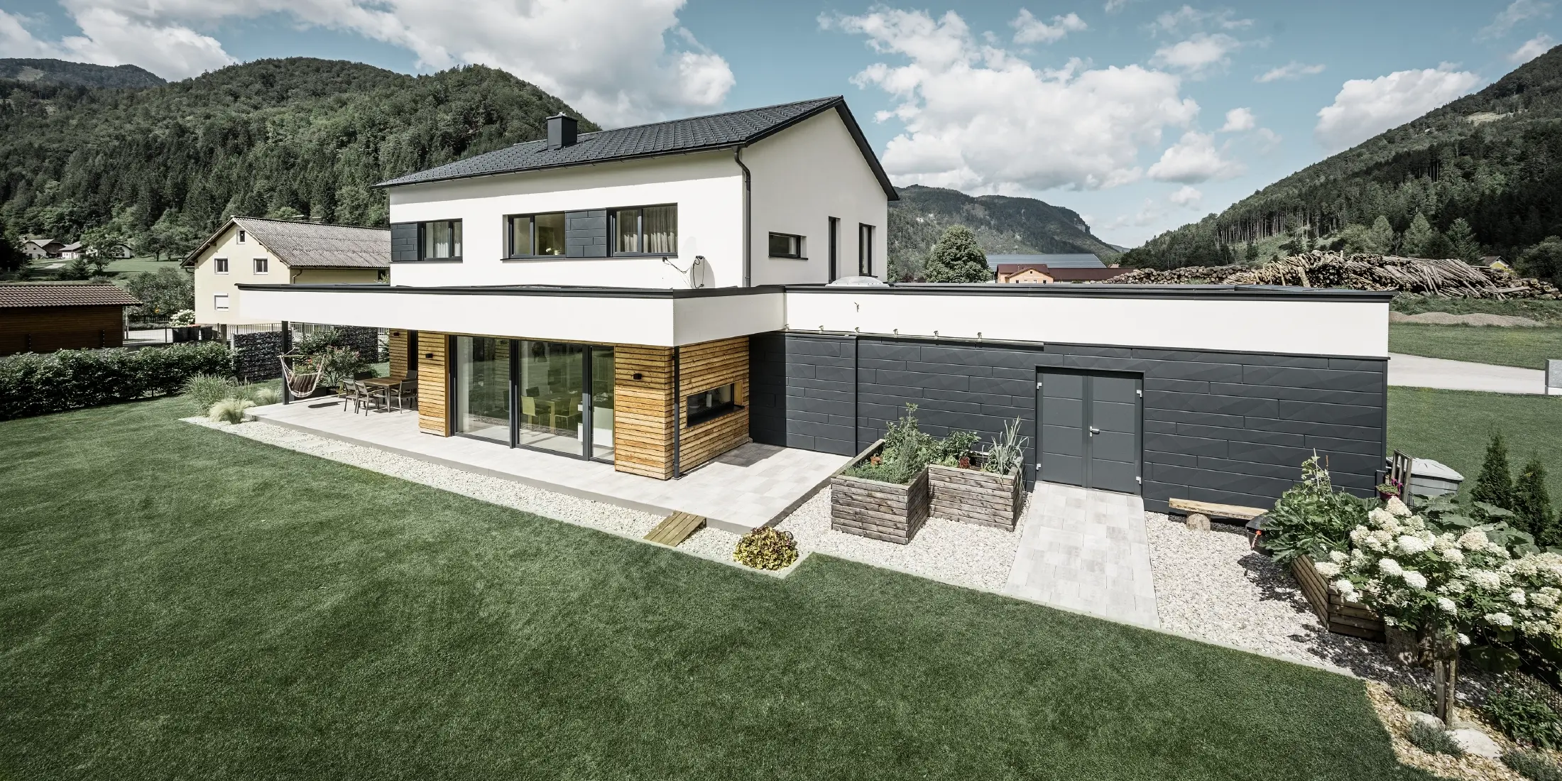 Moderný rodinný dom s bielou omietnutou fasádou a obkladom PREFA Siding.X v antracitovej farbe P.10, ktorý tvorí výrazný kontrast k prírodnému prostrediu. Krytá Falcovanou škridlou PREFA, odvodnenie strechy závesným žľabom PREFA a odtokovou rúrou.