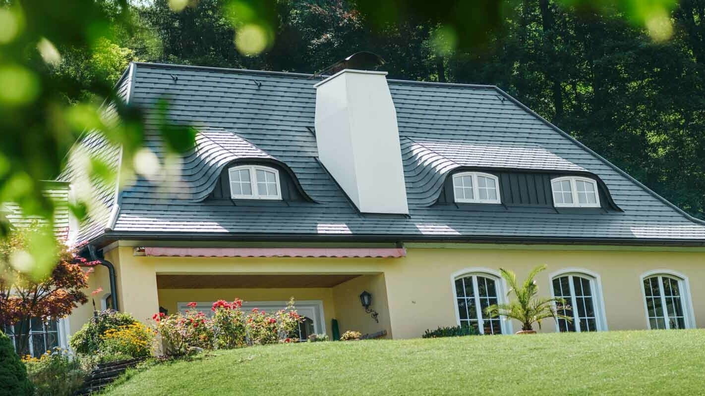 Strecha domu s veľkým komínom a oblúkovým vikierom po rekonštrukcii s použitím PREFA falcovaných šindľov 