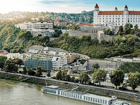 Výhľad na štvrť Vydrica v Bratislave s panorámou hradu a Dunaja: Viacvrstvová architektúra zaujme zelenými strechami, strmými sedlovými strechami a precízne integrovanými strešnými oknami, ktoré vytvárajú výrazné vizuálne akcenty. Hliníkové strešné šindle PREFA vo farbách prefa biela, pieskovo hnedá a bronzová prepájajú jednotlivé objekty do harmonického celku a citlivo ich začleňujú do historického prostredia. Odolné, ľahké a mimoriadne trvácne riešenia PREFA umožňujú realizáciu komplexných tvarov striech a zároveň vytvárajú udržateľný, bezúdržbový obvodový plášť s vysokou estetickou hodnotou.