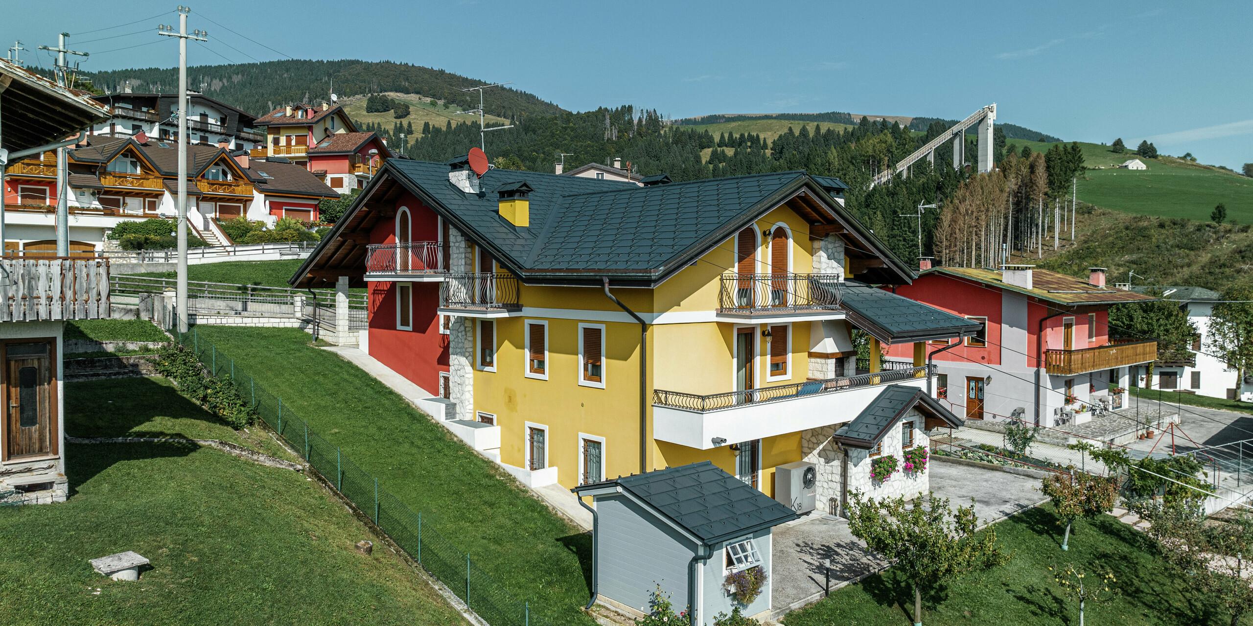 Rodinný dom v Gallio v tradičnom alpskom štýle s jasne žlto-červenou omietkou fasády a prvkami z prírodného kameňa. Strecha z PREFA strešných panelov R.16 v tmavošedej farbe P.10 ponúka najvyššiu odolnosť voči poveternostným vplyvom a dokonale ladí s okolím. Odvodnenie strechy pomocou PREFA odvodňovacieho žľabu a zvodu – tiež v tmavošedej farbe P.10 – zaisťuje spoľahlivú ochranu. Obklopený zelenými lúkami a s výhľadom na hory spája tento dom estetiku, dlhú životnosť a funkčnosť. V pozadí je vidieť známy skokanský mostík v tejto oblasti.