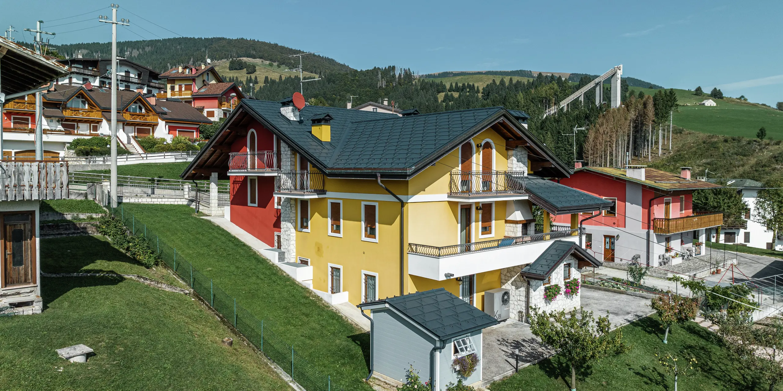 Rodinný dom v Gallio v tradičnom alpskom štýle s jasne žlto-červenou omietkou fasády a prvkami z prírodného kameňa. Strecha z PREFA strešných panelov R.16 v tmavošedej farbe P.10 ponúka najvyššiu odolnosť voči poveternostným vplyvom a dokonale ladí s okolím. Odvodnenie strechy pomocou PREFA odvodňovacieho žľabu a zvodu – tiež v tmavošedej farbe P.10 – zaisťuje spoľahlivú ochranu. Obklopený zelenými lúkami a s výhľadom na hory spája tento dom estetiku, dlhú životnosť a funkčnosť. V pozadí je vidieť známy skokanský mostík v tejto oblasti.