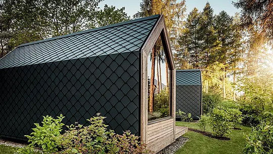 Minimalistický domček Secret Koi Garden pri jazere Natterer See je zasadený do zelene a obklopuje ho teplé večerné svetlo. Strecha a fasáda sú kompletne obložené malými hliníkovými strešnými a fasádnymi šablónami PREFA v odtieni P.10 antracit, ktorých presná štruktúra pripomínajúca rybiu kožu vytvára výrazné akcenty. Odolný a poveternostne odolný hliník PREFA spoľahlivo chráni architektúru a dokonale ladí s drevom, sklom a okolitou prírodou.