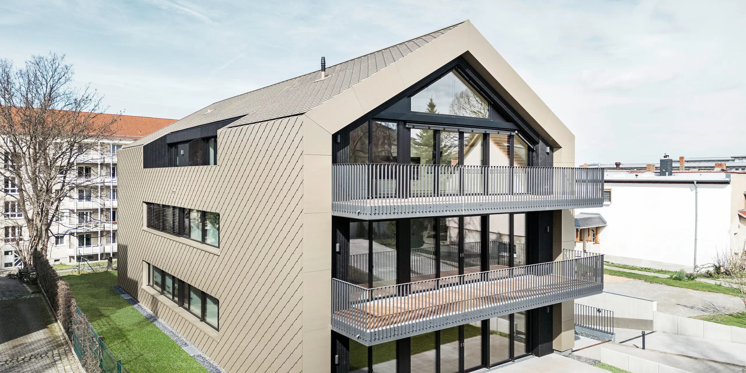 Pohľad do záhrady moderného bytového domu na ulici Chemnitzer Strasse v Drážďanoch. Udržiavaná záhrada leží pred pôsobivou architektúrou. Fasáda a strecha budovy sú obložené odolnou hliníkovou šablónou PREFA 29 × 29 v bronzovej farbe, ktorá dodáva budove elegantný a moderný vzhľad. Na obrázku sú vidieť veľkorysé balkóny, veľké okenné priečelia a harmonické začlenenie budovy do okolia.