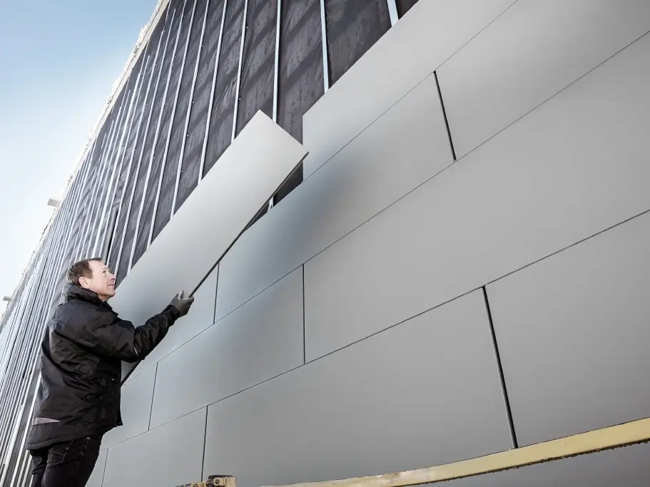 Fotografie čiastočne dokončenej fasády, ktorá je obložená hliníkovými lamelami PREFA Siding v šírke 500. Klampiar montuje ďalší fasádny panel. Fotografia je nasnímaná zospodu. 