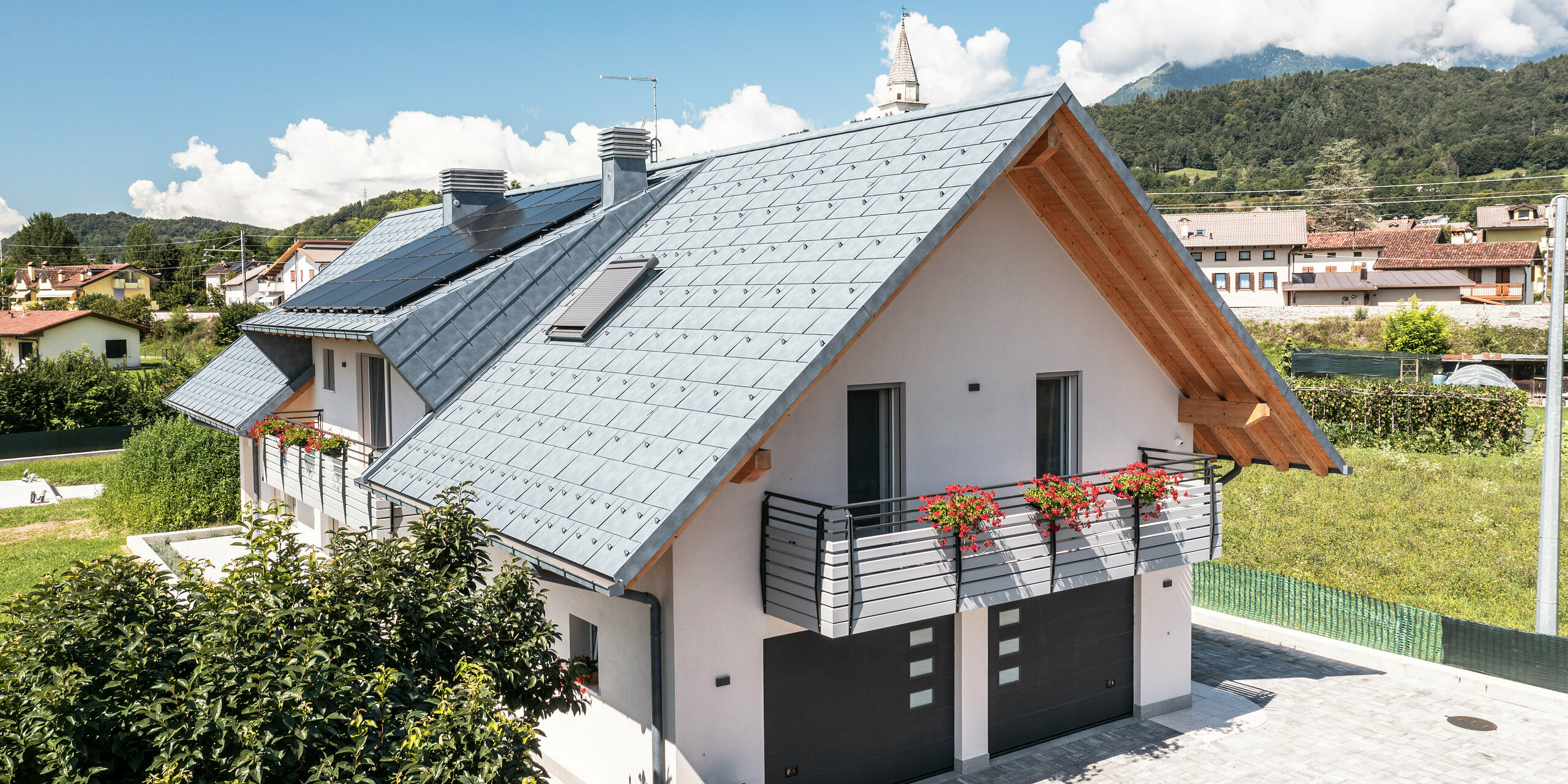 Moderný rodinný dom v talianskom Bellune, charakteristický strešným systémom PREFA vo farbe antracitová v prevedení strešného panelu R.16, ktorý opticky ladí s pohľadovými drevenými trámami štítu. Starostlivo navrhnutý vonkajší priestor s kvitnúcimi balkónmi a dobre udržiavaným živým plotom dodáva nehnuteľnosti prívetivý šmrnc v pokojnej dedinke s kulisou Dolomitov. Pod strechou sú dva vjazdy do garáže. Farba garážovej brány ladí s hliníkovými prvkami a dodáva nehnuteľnosti celkový harmonický vzhľad.