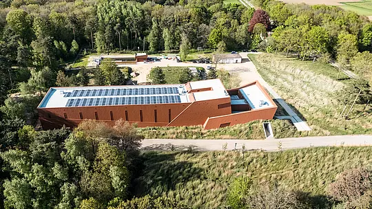 Letecký pohľad na čerpaciu stanicu pitnej vody WML v Lottume ako výraznú infraštruktúrnu stavbu, harmonicky zasadenú do zelenej krajiny lesov a lúk. Podlhovastá budova s výraznou hliníkovou fasádou PREFA Siding.X v teplom vzhľade hrdze pôsobí ako sochársky orientačný bod v teréne. Pozornosť upútavajú jasne štruktúrovaný tvar budovy, integrované fotovoltaické moduly na streche, ako aj jemne fazetovaný povrch fasády. Odolné, ľahké a recyklovateľné hliníkové panely PREFA presviedčajú vysokou odolnosťou voči poveternostným vplyvom, trvalo udržateľnou efektívnosťou a fasádou s nízkymi nárokmi na údržbu.