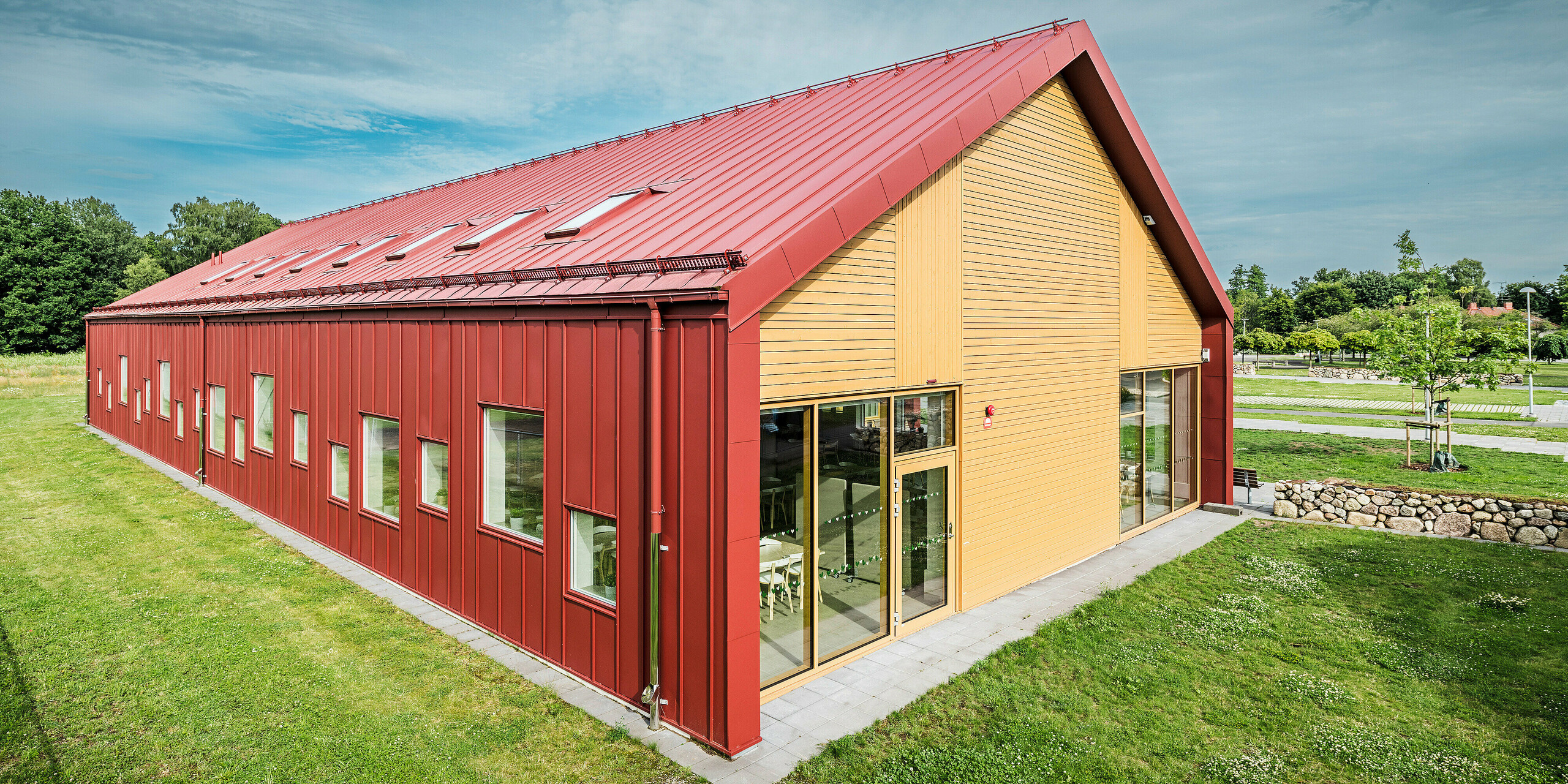 Bočný pohľad na školskú jedáleň Restaurang Gröna Hjärtat v Broby, Švédsko. Na obrázku je zobrazený štít budovy s pútavou kombináciou hliníkového obkladu PREFALZ v tmavočervenej farbe a teplej drevenej fasády. Čisté, moderné línie architektúry a farebné kontrasty medzi červeným obkladom so stojatou drážkou a prírodným drevom zdôrazňujú inovatívny dizajn budovy. Okná a vstupné dvere na tejto strane prepúšťajú do miestností dostatok prirodzeného svetla a prispievajú k príjemnej atmosfére. Odolná konštrukcia z kvalitného plechu zdôrazňuje kvalitu a udržateľnosť budovy.