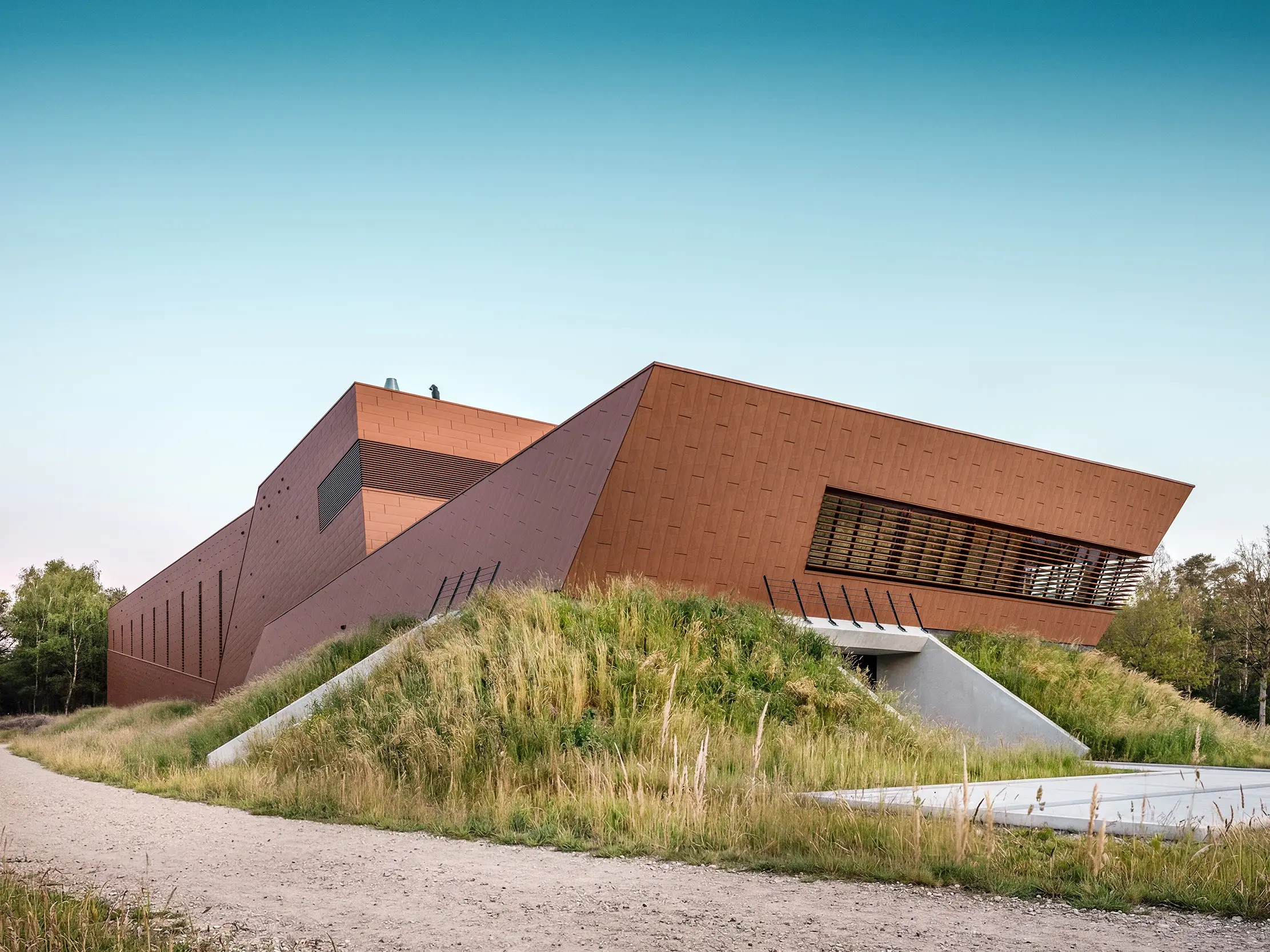 Architektonicky výrazná čerpacia stanica pitnej vody WML v Lottume so sochársky tvarovanými stavebnými telami, zasadená do prírodnej krajiny s násypmi. Výrazná hliníková fasáda PREFA Siding.X v teplom vzhľade hrdze priťahuje všetky pohľady svojím fazetovaným, živým povrchom a pôsobivo hrá so svetlom a tieňom. Vodorovné lamely a presné línie zdôrazňujú dynamickú geometriu. Ľahké, trvanlivé a úplne recyklovateľné hliníkové panely PREFA sú zárukou najvyššej odolnosti voči poveternostným vplyvom, udržateľného stavebného riešenia a obvodového plášťa budovy s nízkymi nárokmi na údržbu.