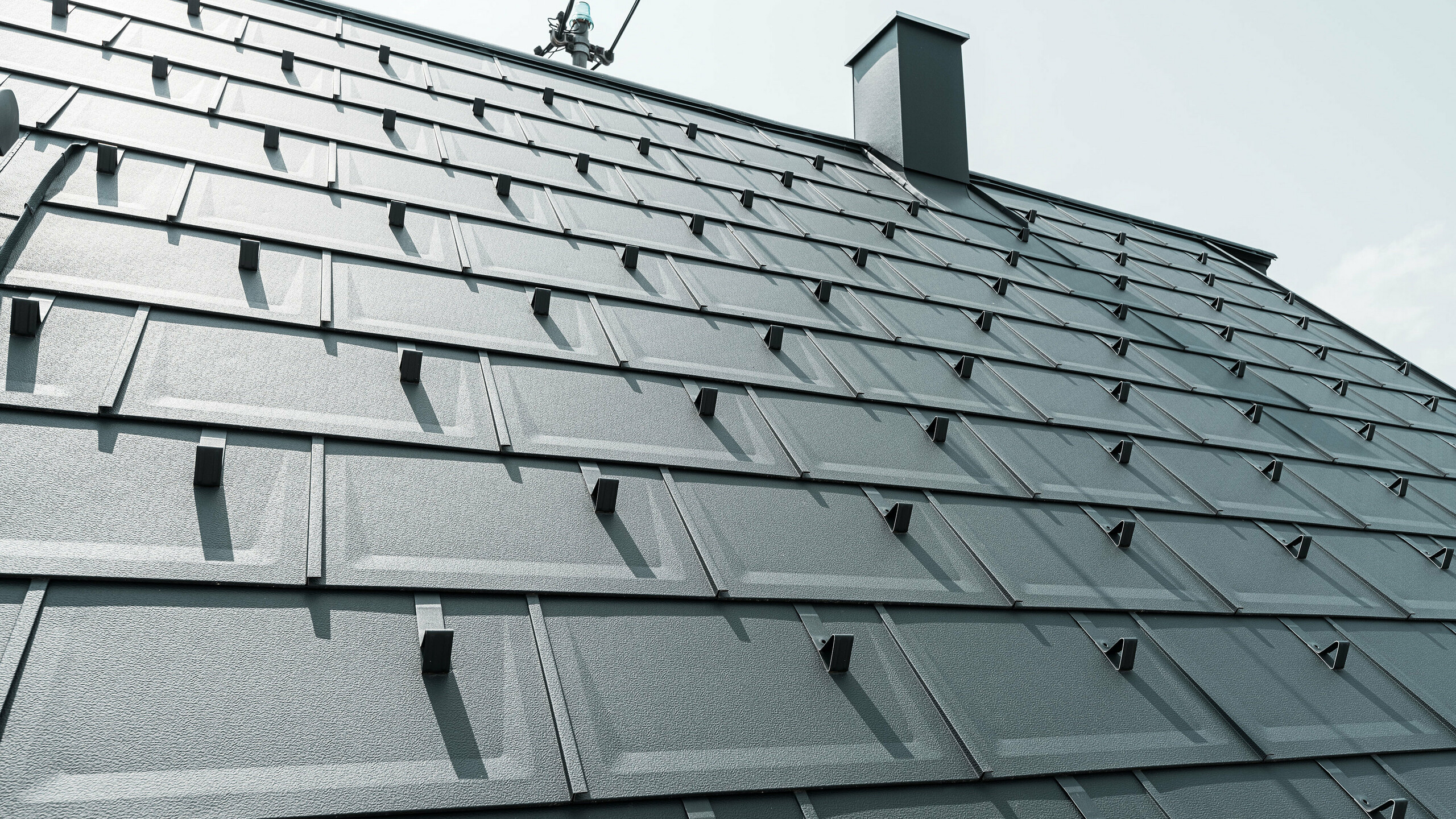 Detailný pohľad na strešný panel PREFA R.16 v farbe P.10 antracit. Na streche sú viditeľné snehové zábrany PREFA a komín.