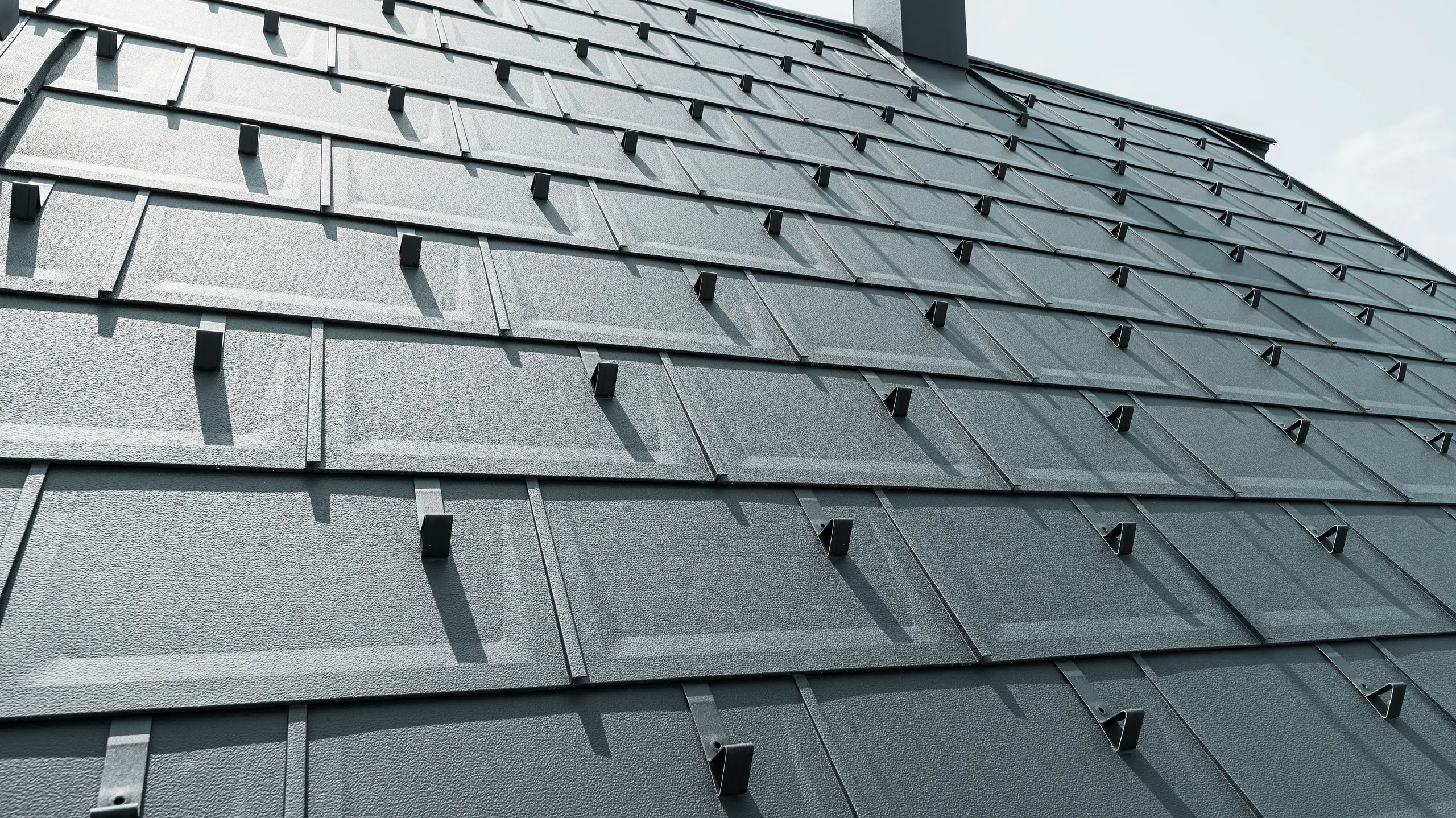 Detailný pohľad na strešný panel PREFA R.16 v farbe P.10 antracit. Na streche sú viditeľné snehové zábrany PREFA a komín.