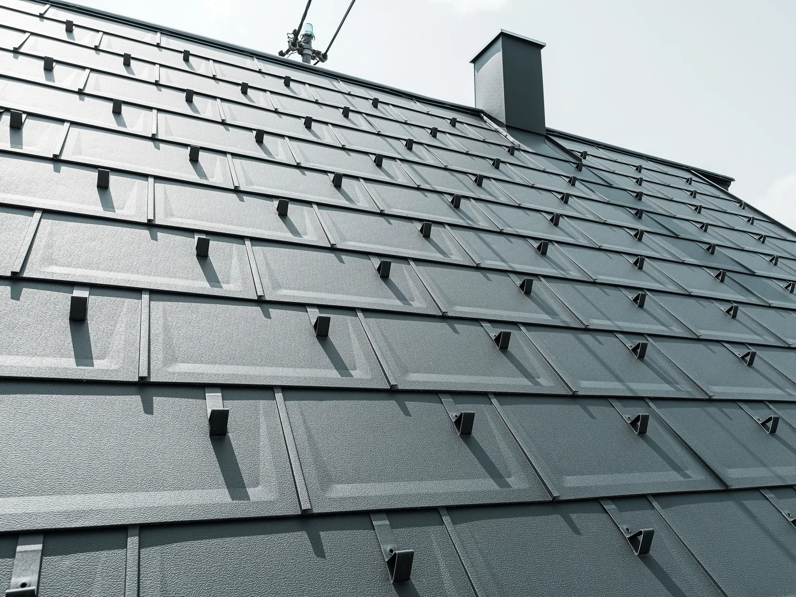 Detailný pohľad na strešný panel PREFA R.16 v farbe P.10 antracit. Na streche sú viditeľné snehové zábrany PREFA a komín.