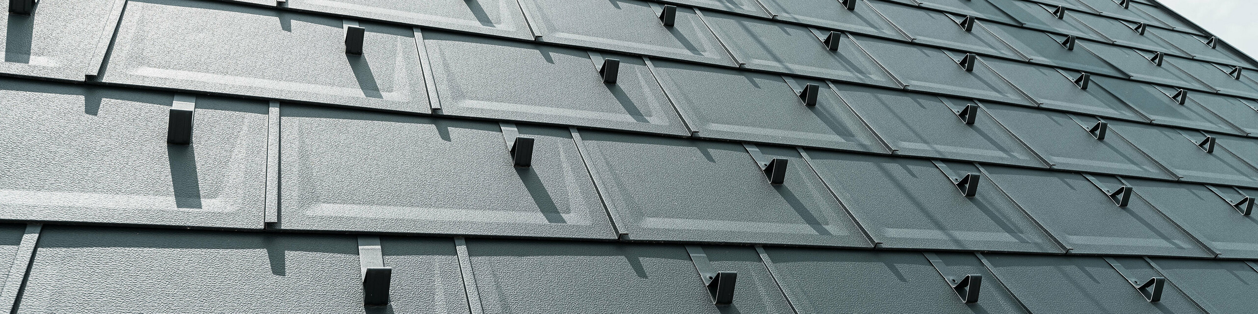 Detailný pohľad na strešný panel PREFA R.16 v farbe P.10 antracit. Na streche sú viditeľné snehové zábrany PREFA a komín.