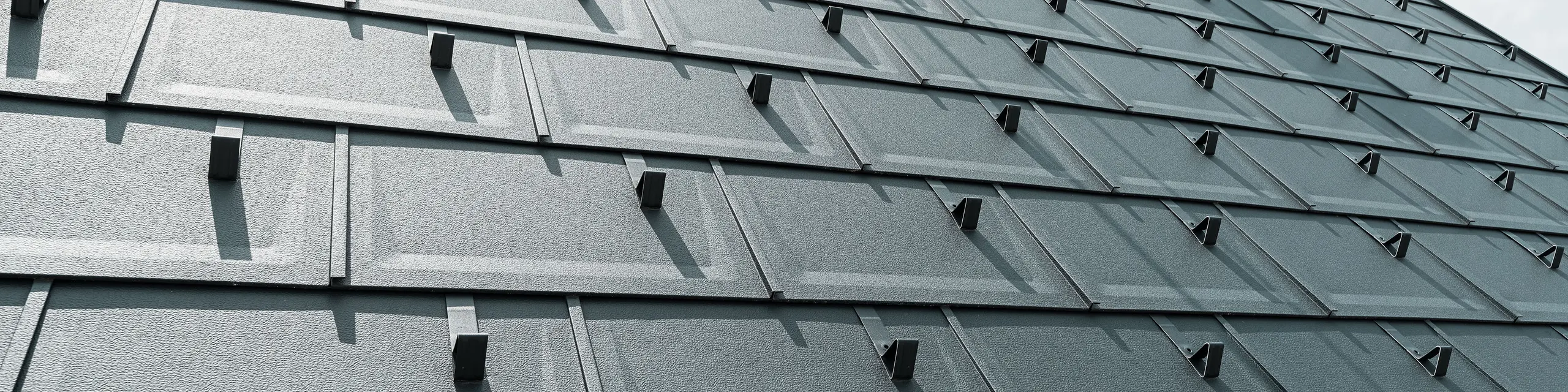 Detailný pohľad na strešný panel PREFA R.16 v farbe P.10 antracit. Na streche sú viditeľné snehové zábrany PREFA a komín.