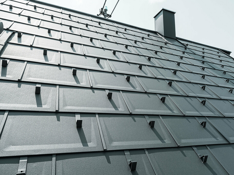 Detailný pohľad na strešný panel PREFA R.16 v farbe P.10 antracit. Na streche sú viditeľné snehové zábrany PREFA a komín.