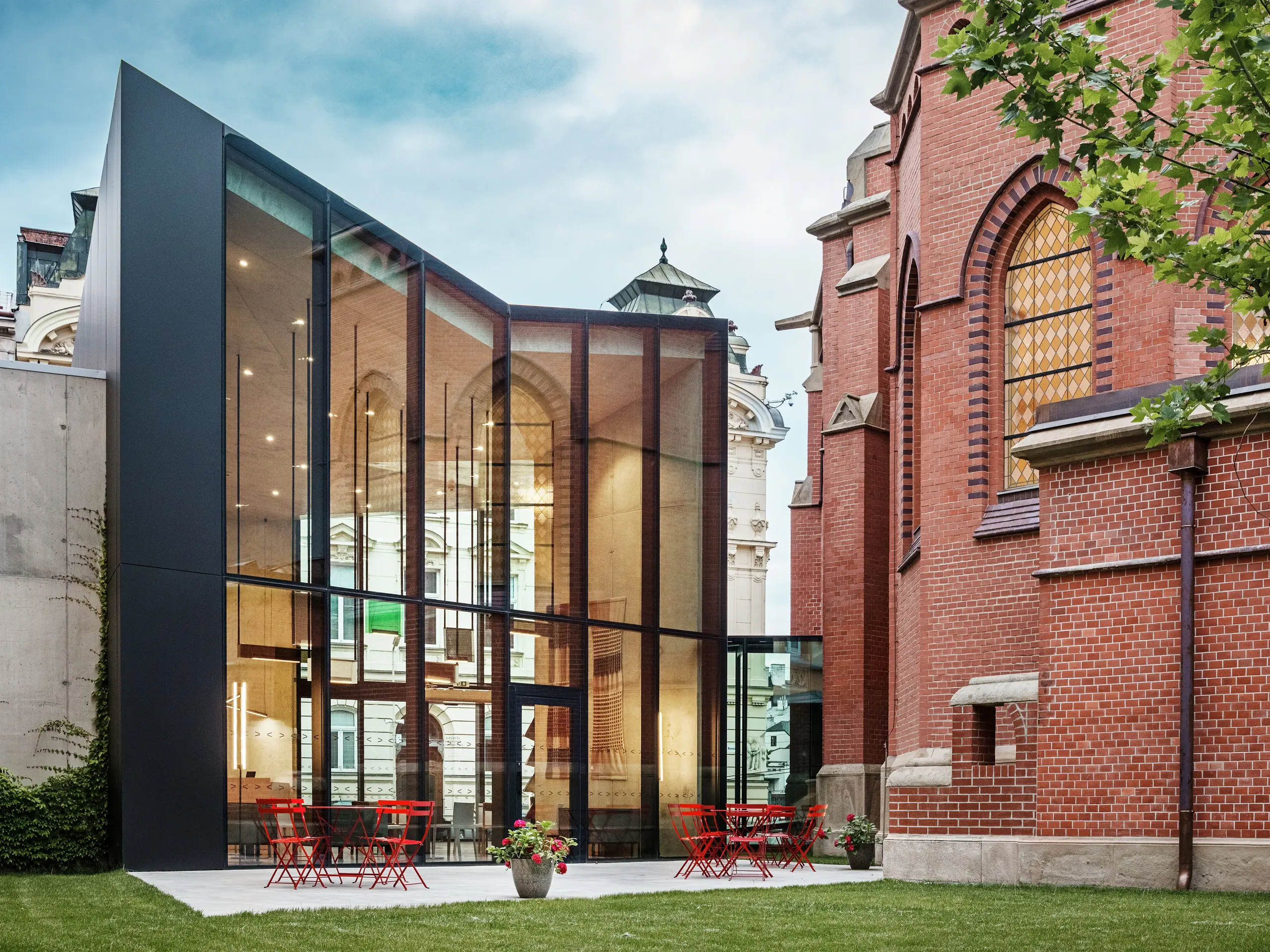 Obrázok ukazuje fascinujúcu novostavbu v Olomouci, ktorá spája historický Červený kostol s modernou knižnicou. Výrazná čierna fasáda z hliníkových kompozitných dosiek PREFABOND vytvára silný kontrast s teplými, červenými tehlami kostola. Veľké sklenené plochy ponúkajú jasný pohľad do vnútra budovy, kde sa rozprestiera moderná, svetlom preplnená atmosféra. Na terase vás červené stoly a stoličky pozývajú k oddychu a vytvárajú harmonické prepojenie medzi historickou budovou a súčasnou architektúrou. Toto elegantné spojenie starého a nového ukazuje výhody dosiek PREFABOND – nielenže zaisťujú moderný estetický dojem, ale ponúkajú aj vynikajúcu životnosť a funkčnosť.