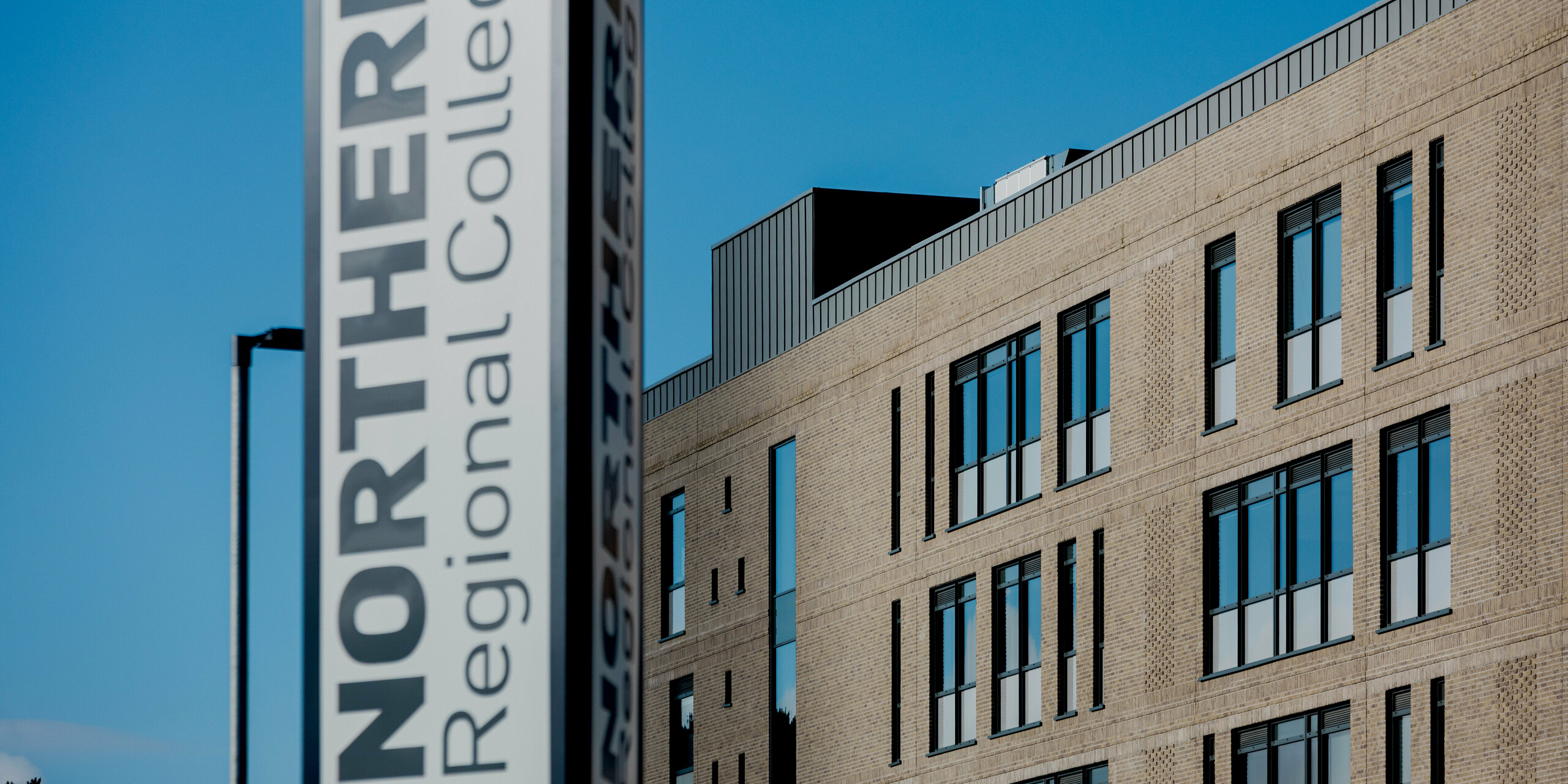 Im Vordergrund steht die vertikale Stele mit der Aufschrift „Northern Regional College“, dahinter entfaltet sich die Klinkerfassade des Campus in Ballymena unter klarem blauem Himmel. Entlang der Dachkante setzen PREFALZ Aluminium-Stehfalzbahnen in Patinagrau einen präzisen, horizontalen Abschluss. Die schmalen Fensterformate strukturieren die Fassade rhythmisch und betonen die lineare Architektur. PREFA Aluminium verleiht dem Bildungsbau eine langlebige, wartungsarme und zeitlos moderne Ausstrahlung.