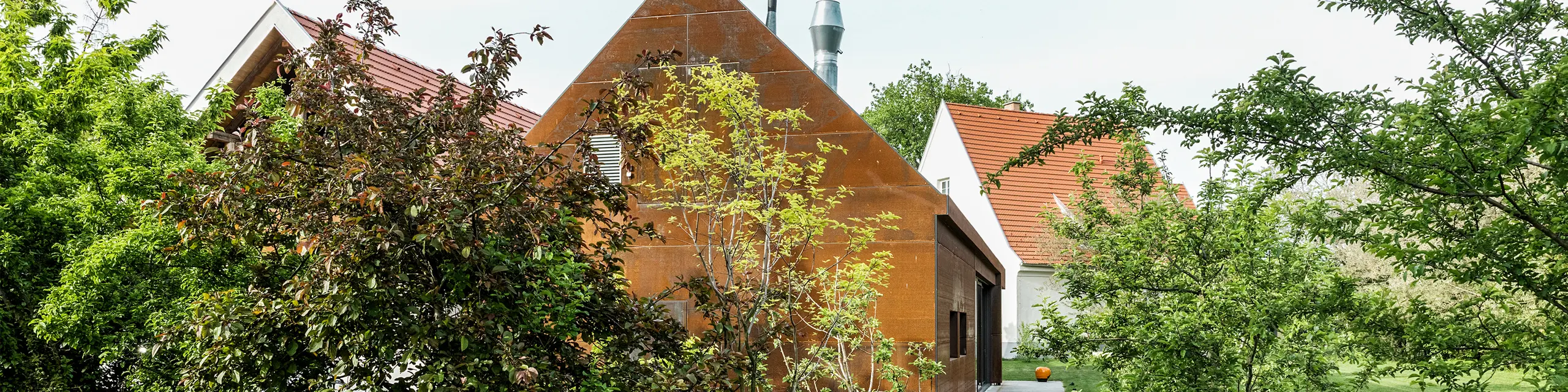 Rezidencia Velich v Seewinkel, Burgenland: pohľad na modernú budovu obklopenú bujnou vegetáciou. Rezidencia slúži ako miesto odborného dialógu PREFARENZEN 2025, v rámci ktorého architekti a odborníci z PREFA vyberajú najzaujímavejšie architektonické projekty. V popredí je vidieť štýlovú kovovú stenu hrdzavej farby s nápisom "RESIDENZ VELICH". Architektúra budovy spája tradičné a moderné prvky a harmonicky zapadá do okolitej prírody.