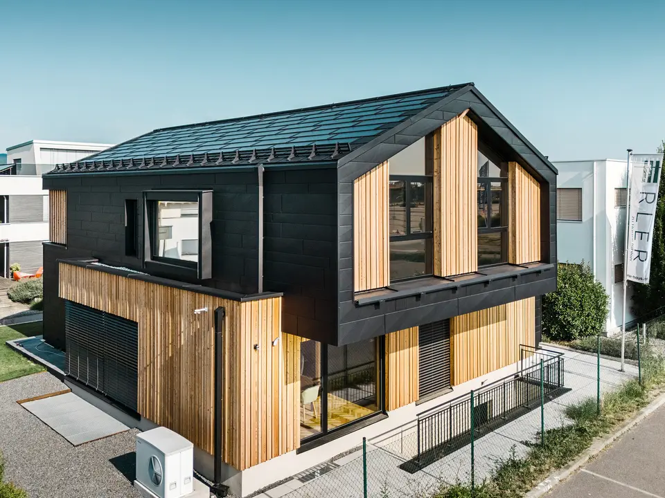 Predná časť modelového domu Autark v centre montovaných domov Blaue Lagune, Vösendorf (AT). Strecha je pokrytá integrovanými solárnymi panelmi PREFA a strešnými panelmi R.16 s kvalitným náterom P.10. Fasádu tvorí kombinácia vertikálnych drevených prvkov a PREFA Siding.X vo farbe P.10 čierna, ktorá dáva budove moderný a udržateľný charakter. Vlajka Erler Bau GmbH veje na pravom okraji obrázku.