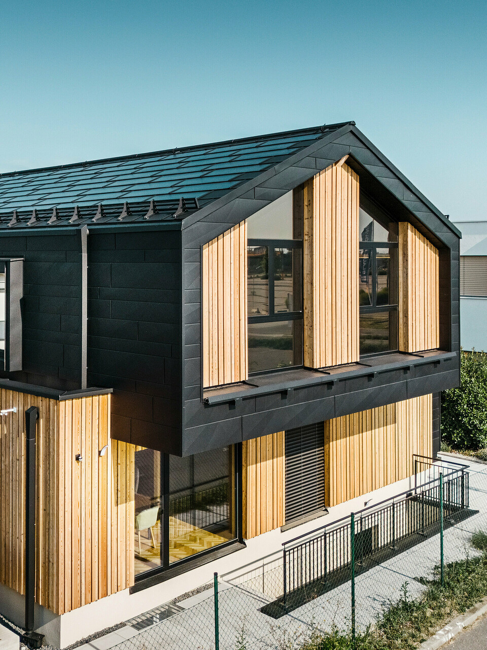 Predná časť modelového domu Autark v centre montovaných domov Blaue Lagune, Vösendorf (AT). Strecha je pokrytá integrovanými solárnymi panelmi PREFA a strešnými panelmi R.16 s kvalitným náterom P.10. Fasádu tvorí kombinácia vertikálnych drevených prvkov a PREFA Siding.X vo farbe P.10 čierna, ktorá dáva budove moderný a udržateľný charakter. Vlajka Erler Bau GmbH veje na pravom okraji obrázku.