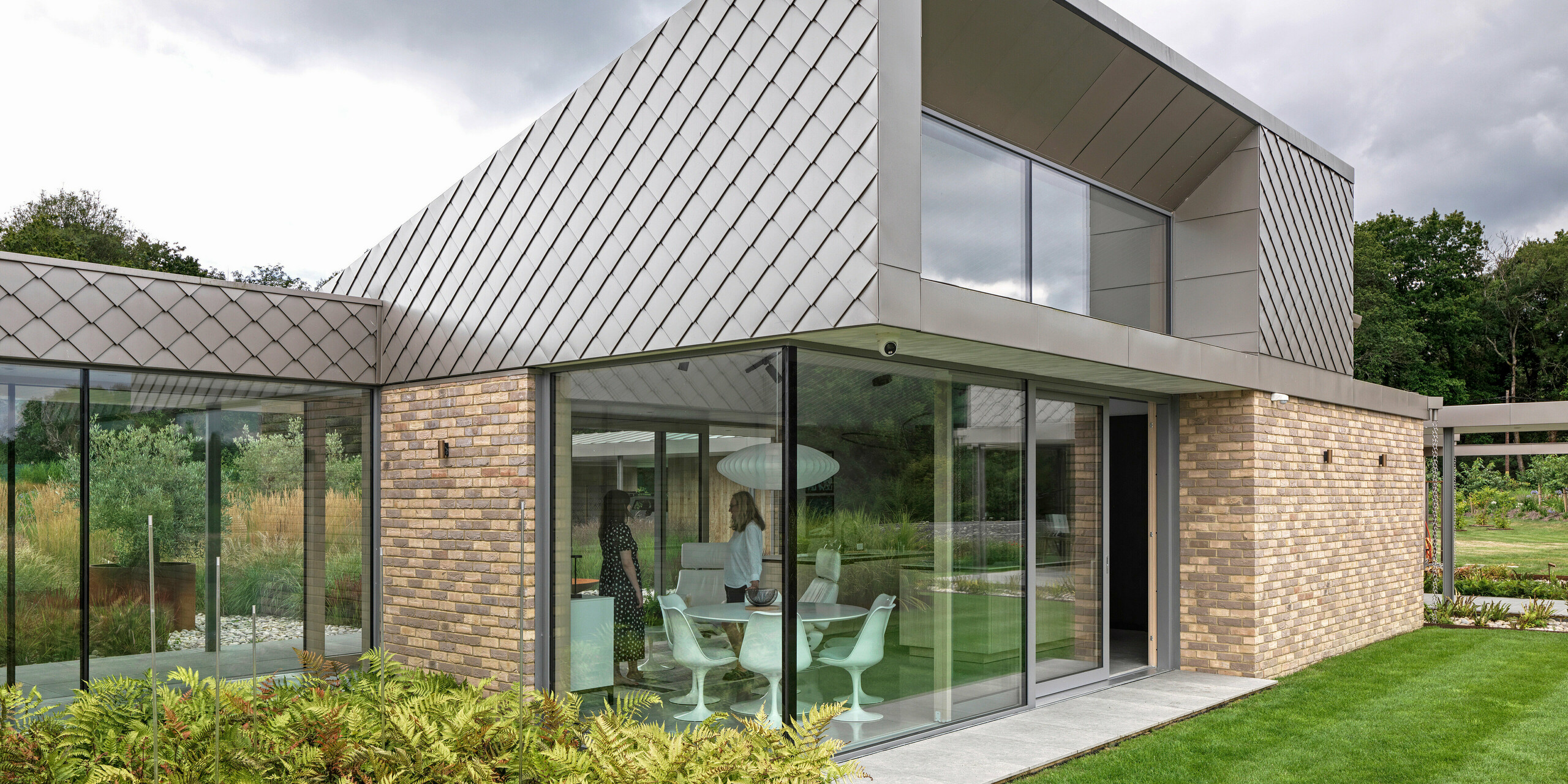 Architektonicky výrazná časť obytného domu v West Sussex s moderným dizajnom fasády z PREFA šablón 20 × 20 v P.10 bronzovej farbe. Strešné plochy sú pokryté hliníkom PREFALZ a svojimi čistými líniami podčiarkujú nadčasový charakter návrhu. Veľkorysé sklenené fasády vytvárajú svetlé obytné priestory a opticky spájajú budovu s udržiavanou záhradou. Vysoko kvalitné produkty PREFA ponúkajú dlhú životnosť a jednotný vzhľad pri minimálnej údržbe.