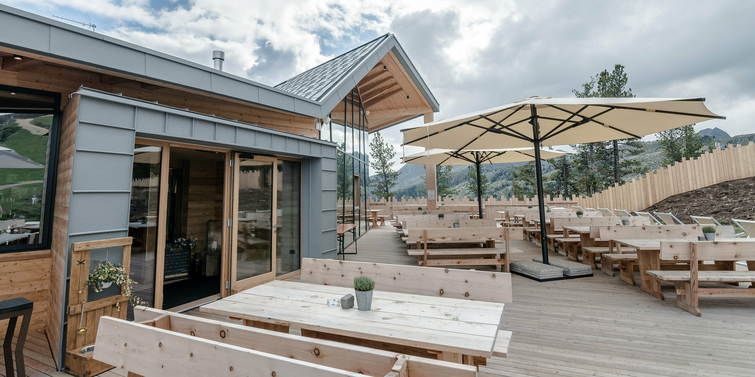 Ristorante Busabelle, obklopená pôsobivou horskou krajinou, má priestrannú terasu s robustnými drevenými stolmi a elegantnými slnečníkmi, ktoré poskytujú príjemné posedenie. Architektúra kombinuje modernú svetlosivú fasádu PREFALZ s prírodnými drevenými prvkami a zužujúcou sa strechou, čo dodáva budove sviežu a príjemnú atmosféru.