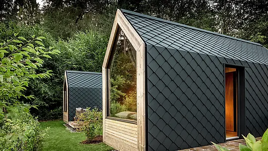 Dva minimalistické minidomy Secret Koi Garden sú zasadené do bujnej zelene pri jazere Natterer See. Fasády a strechy sú kompletne obložené malými hliníkovými šablónami PREFA v odtieni P.10 antracit, čím vytvárajú výraznú štruktúru pripomínajúcu rybiu kožu. Veľké sklenené plochy a teplé drevené detaily vytvárajú zaujímavý kontrast s tmavým hliníkom. Odolný a poveternostne odolný hliník PREFA spoľahlivo chráni a podčiarkuje vysokú kvalitu architektúry glampingu.