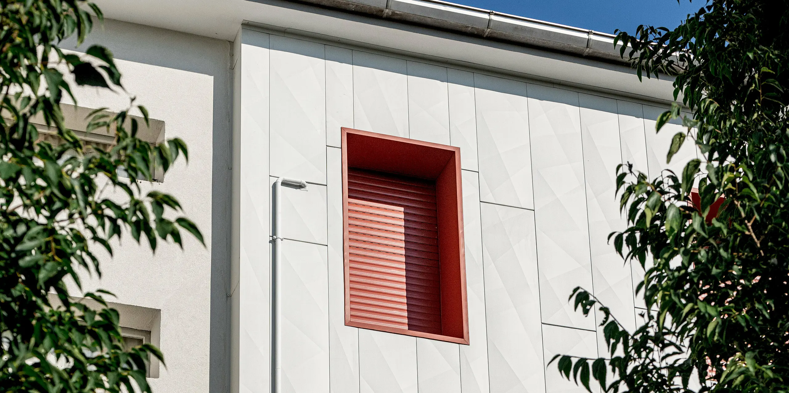 Moderne Wohnanlage Alba B nach der Sanierung mit PREFA Siding.X Aluminiumfassade: Die vertikal verlegten Paneele erzeugen spannende Licht- und Schattenspiele auf der hellen Oberfläche. Markante rote Fensterrahmen setzen starke Kontraste und lenken den Blick gezielt auf die Fassadenstruktur. Umgeben von grünen Baumkronen wirkt das Gebäude lebendig und hochwertig. Die PREFA Aluminiumfassade überzeugt durch Langlebigkeit, Widerstandsfähigkeit und Wartungsarmut und bietet zugleich eine zeitgemäße, architektonisch prägnante Optik.