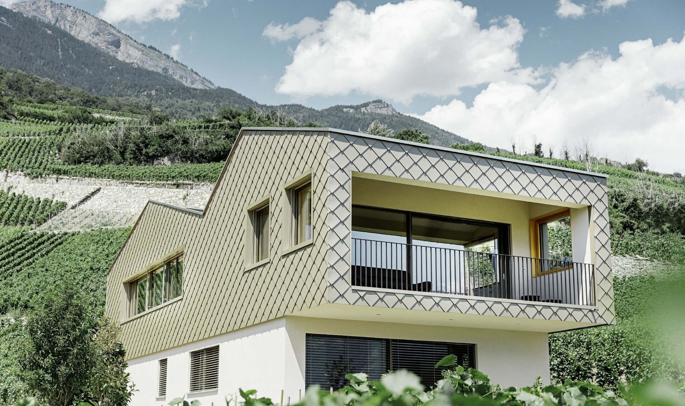 Moderný rodinný dom uprostred vinohradov v údolí Rhônetal so 4 rôznymi strešnými plochami a otvorenou galériou so kosoštvorcovými fasádadnymi šablónami bronzovej farby